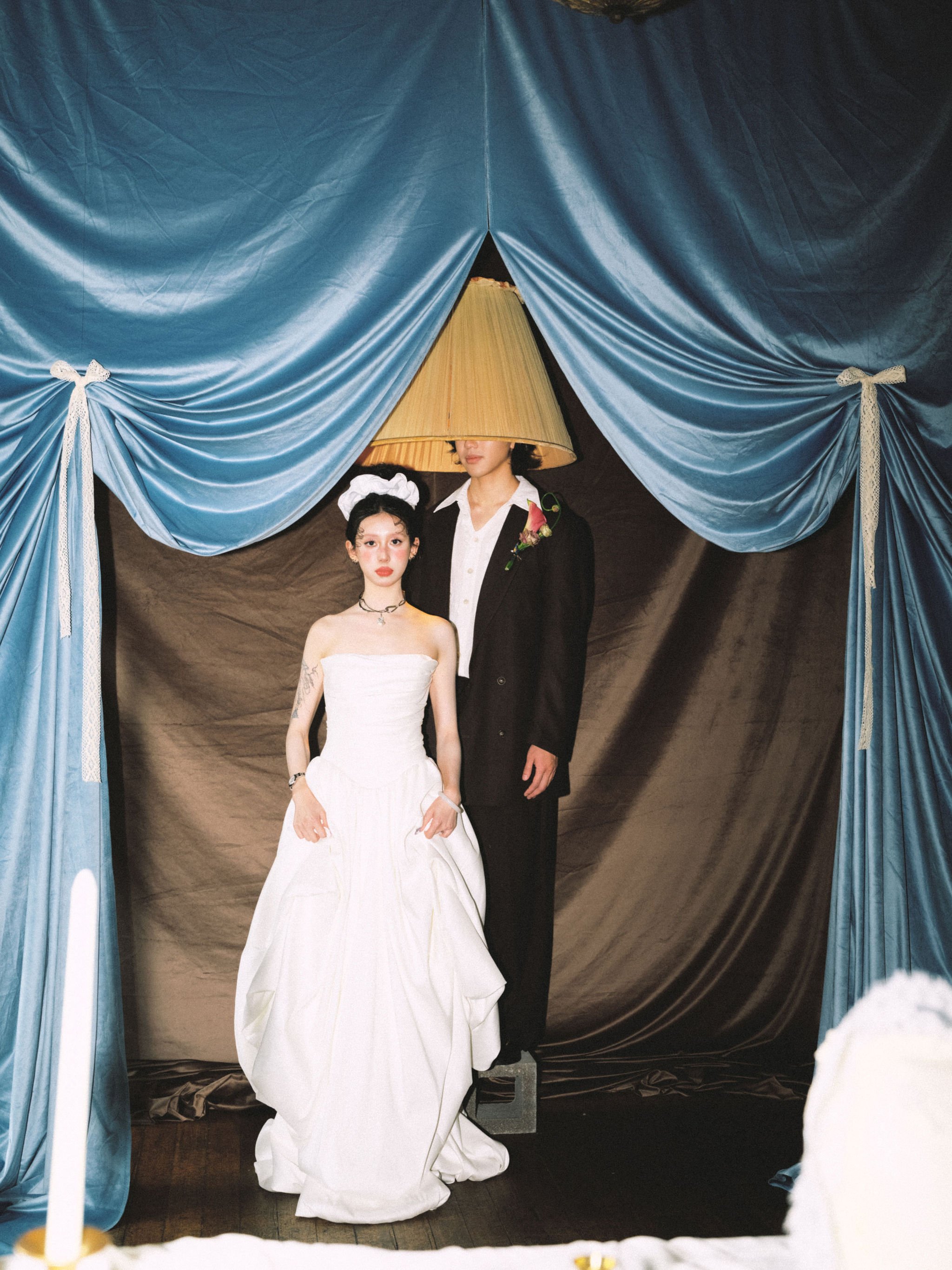 Two women in wedding attire are standing at a ceremony or photoshoot, with blue curtains framing the scene. One woman is wearing a white strapless gown with tattoos on her arm and a choker necklace, and the other woman, whose face is partly covered by a large lampshade, is dressed in a black suit with a boutonniere on the lapel.