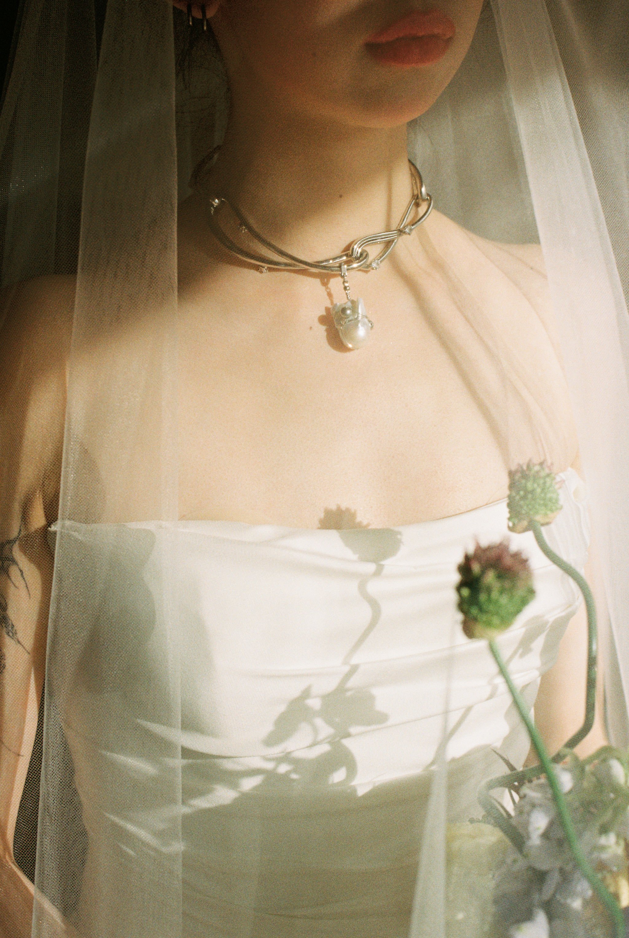 A person with long light-colored hair wearing a white strapless top, standing behind a sheer curtain with floral patterns. They are wearing a chunky silver necklace with a pendant and a partially visible earring. Two stems with green, rounded buds or flowers are in the foreground, casting shadows on the person's chest.