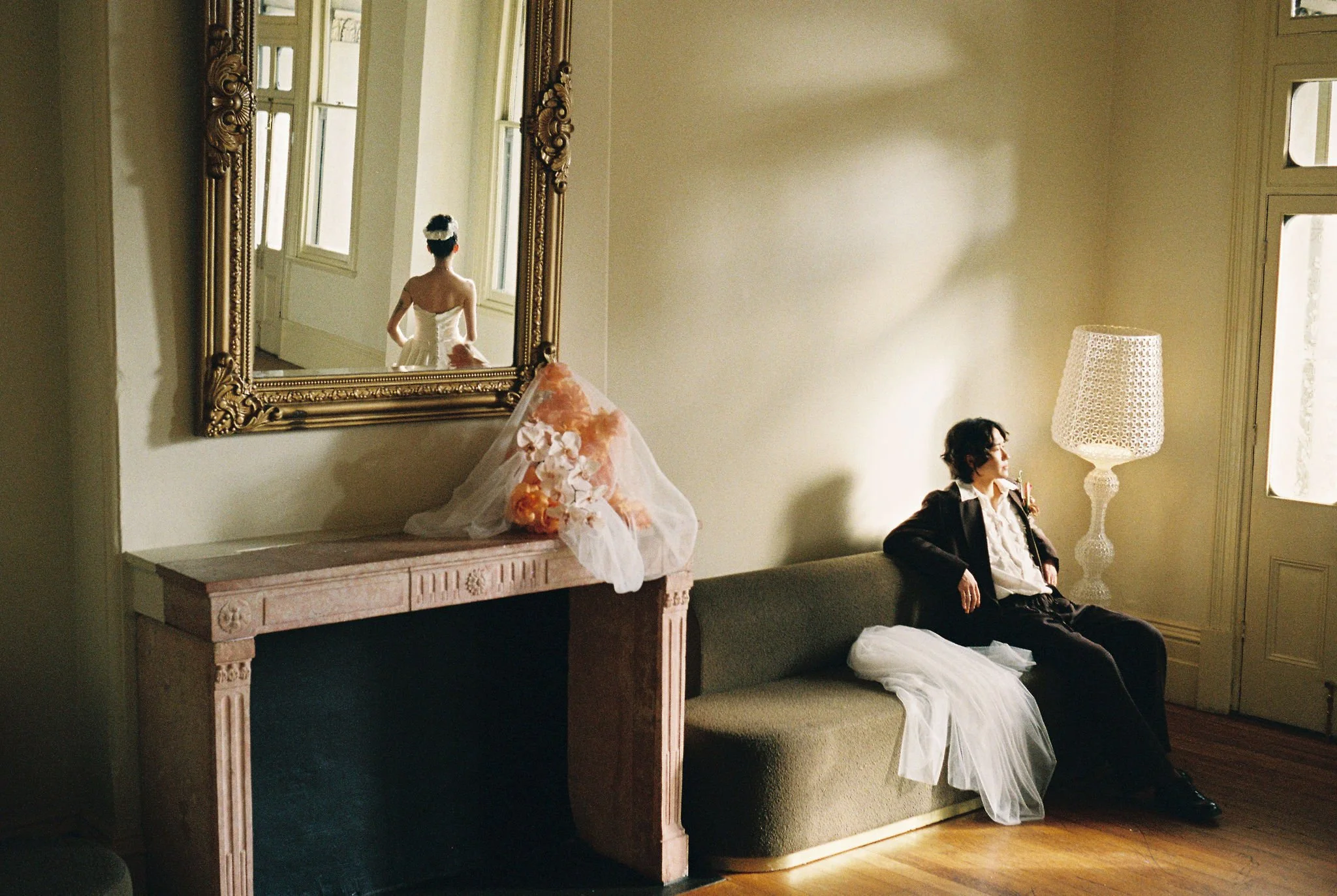 A woman in a wedding dress looking out a window, and a man in a tuxedo sitting on a bench nearby, with a large mirror reflecting the woman's back.