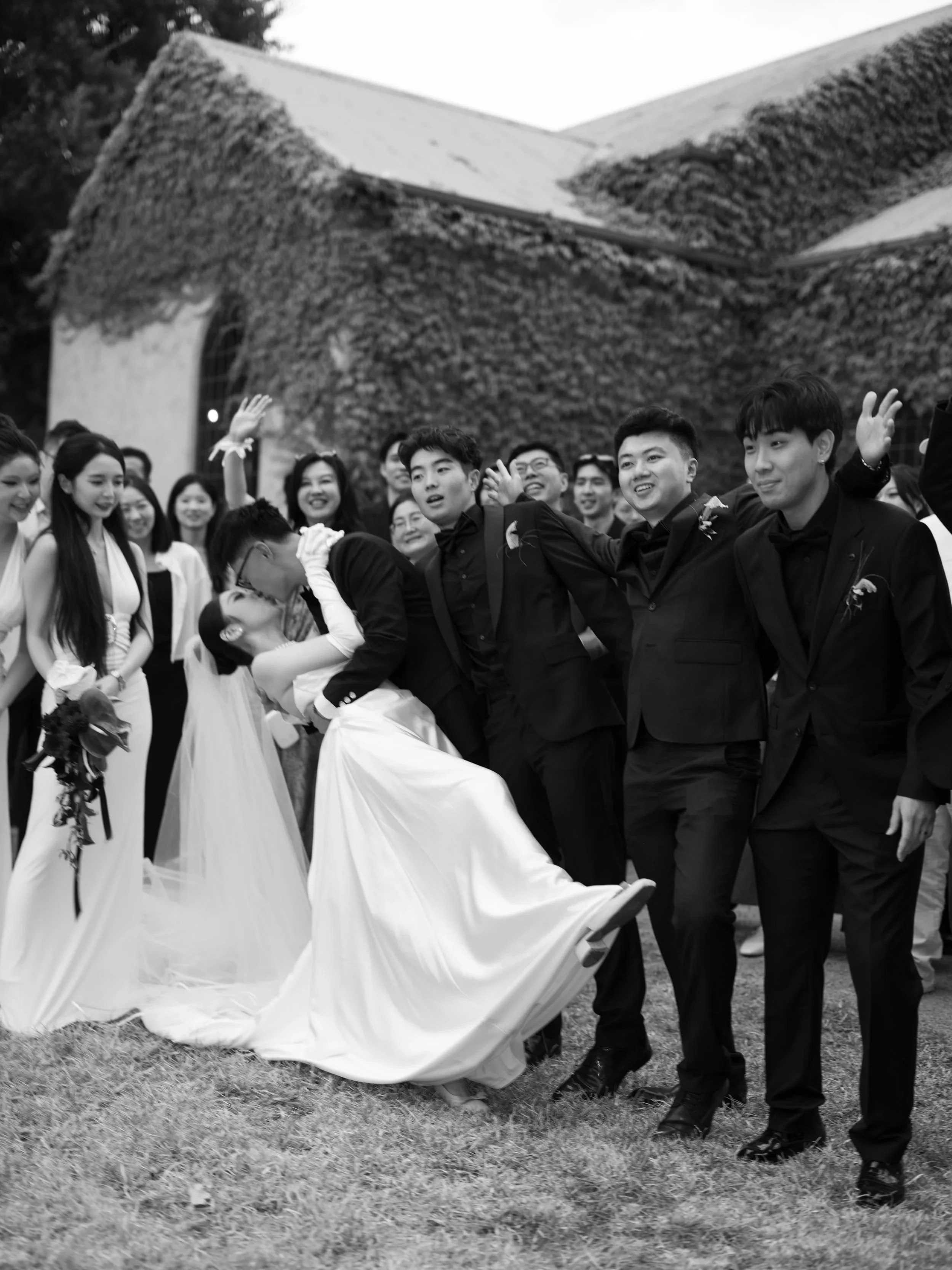 Group of people celebrating, with some dressed in formal attire and one person in a wedding dress, outdoors on grass, with a building covered in ivy in the background.