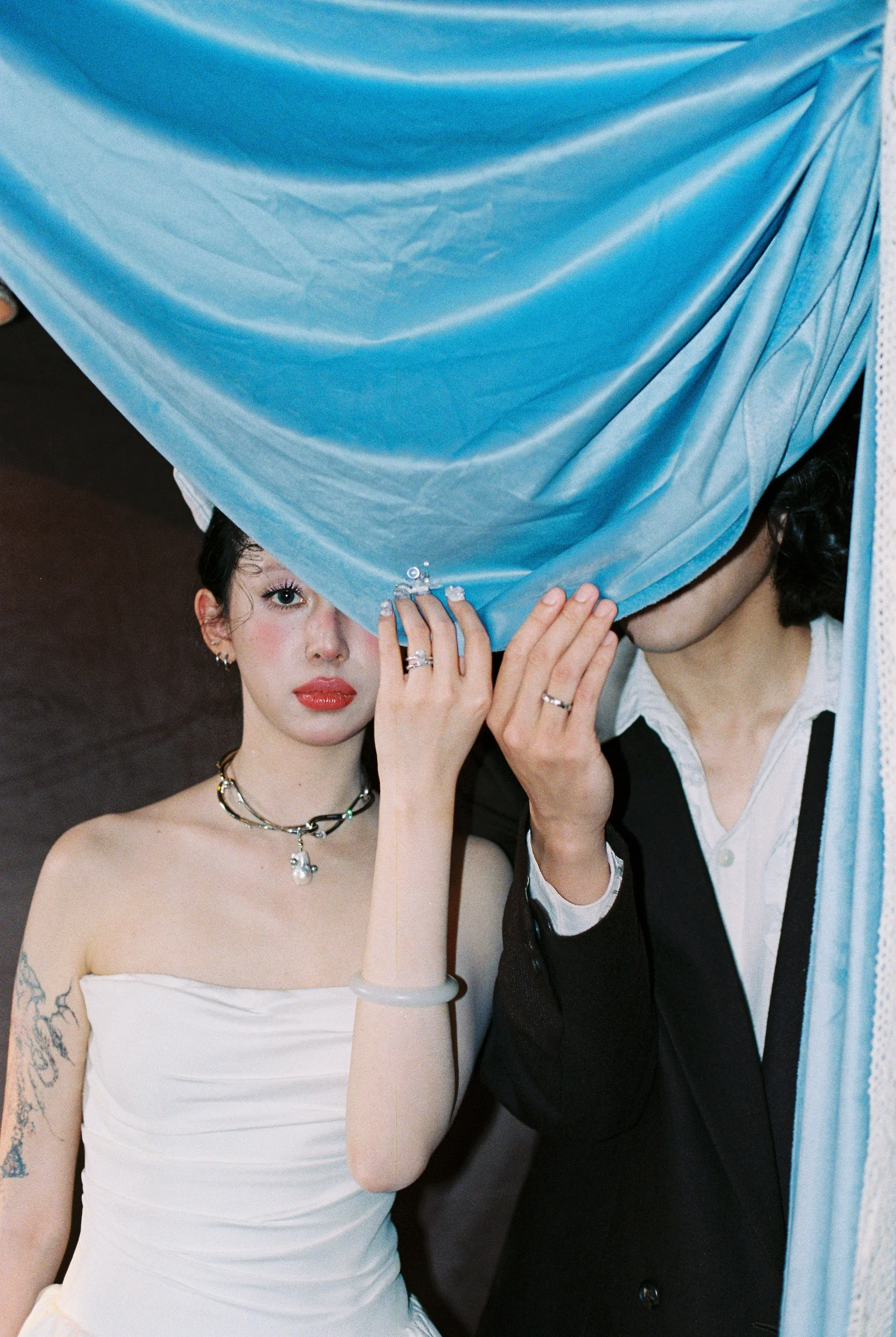 Two women are behind a blue curtain. One woman with dark curly hair, wearing a black and white outfit, is holding the curtain, and the other woman with dark hair, wearing a white strapless top, is looking towards the camera.