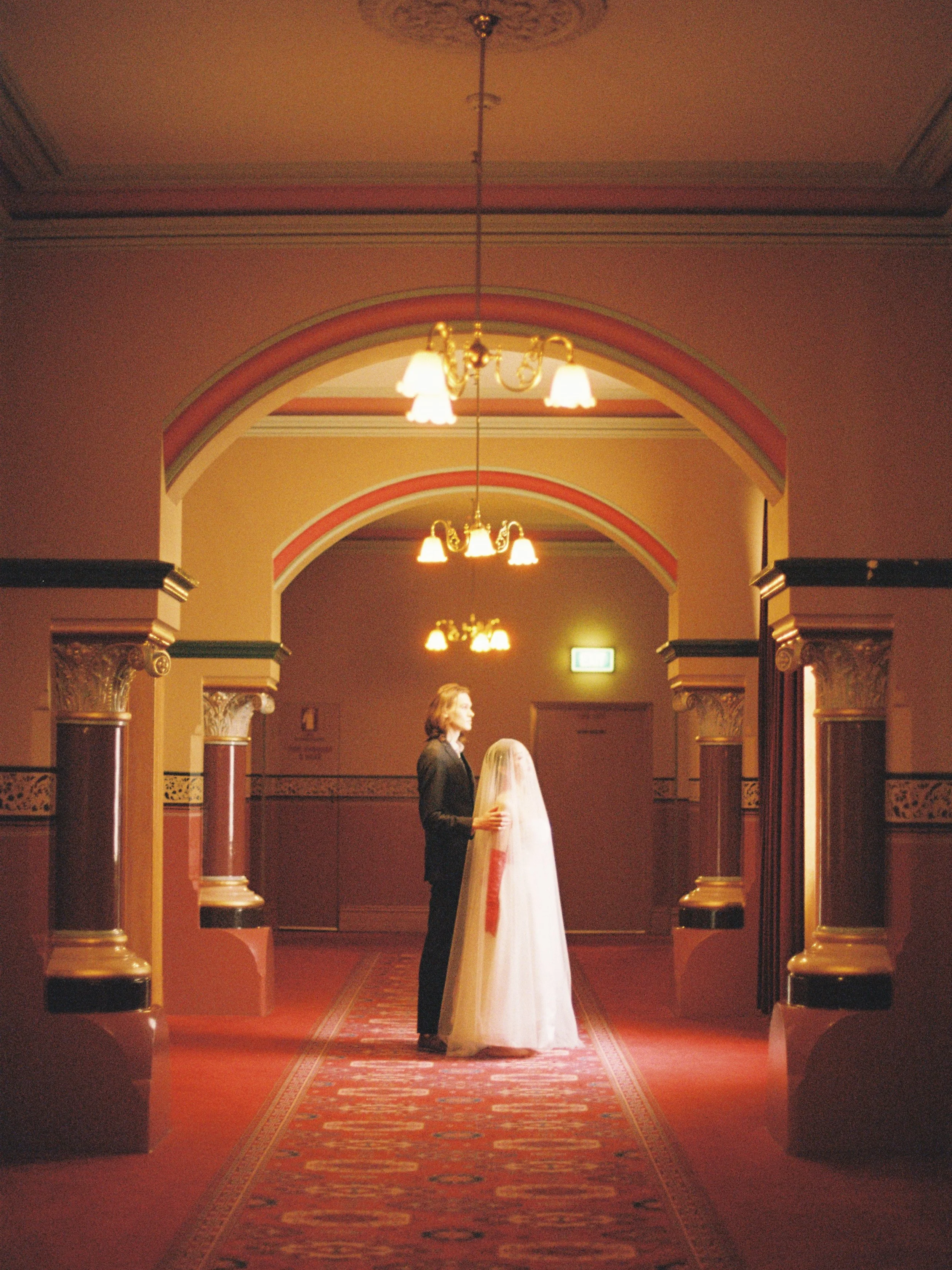 Interior of a hallway with warm lighting, ornate columns, and an arched ceiling. In the center, a man in a black suit stands next to a person in a wedding dress.