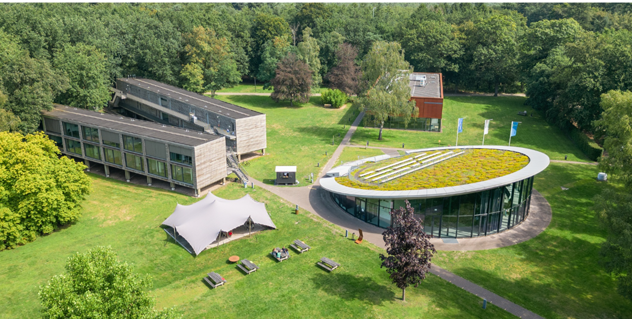 Luchtfoto van een modern gebouw met een ronde structuur, omgeven door groene grasvelden, bomen en een tentenopstelling in een parkachtige omgeving.