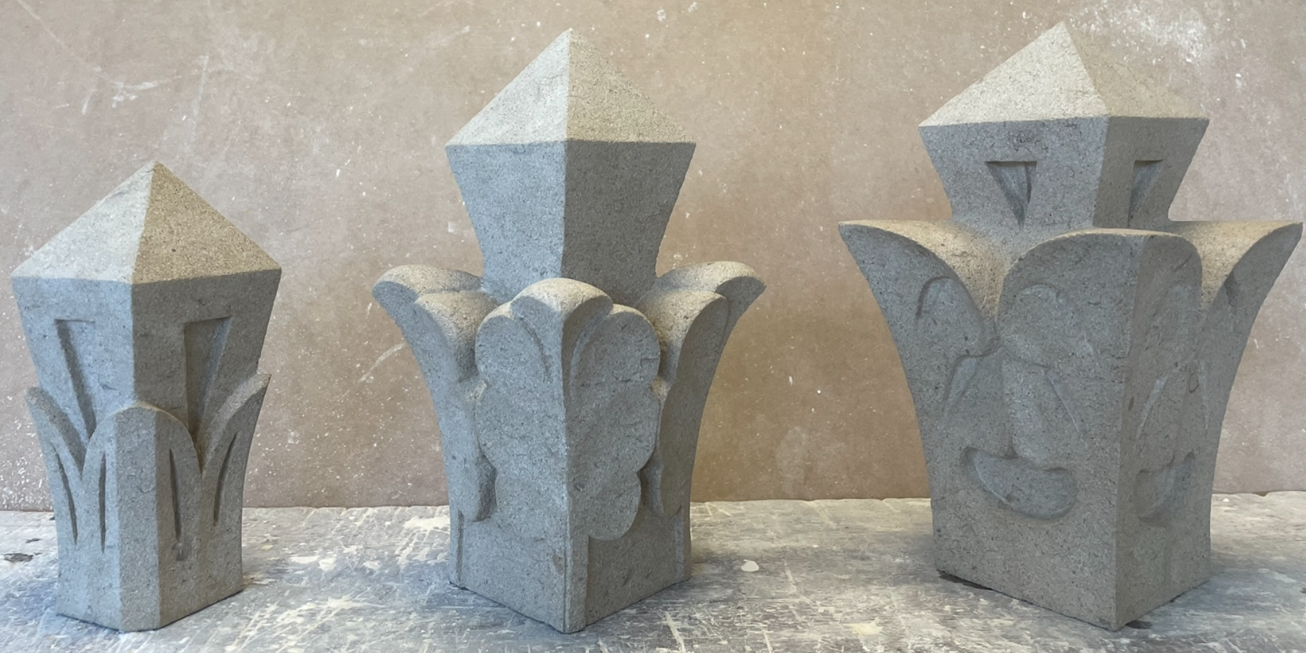 Three decorative stone sculptures resembling stylized flowers or plants with geometric shapes on top, placed on a textured surface against a plain background.