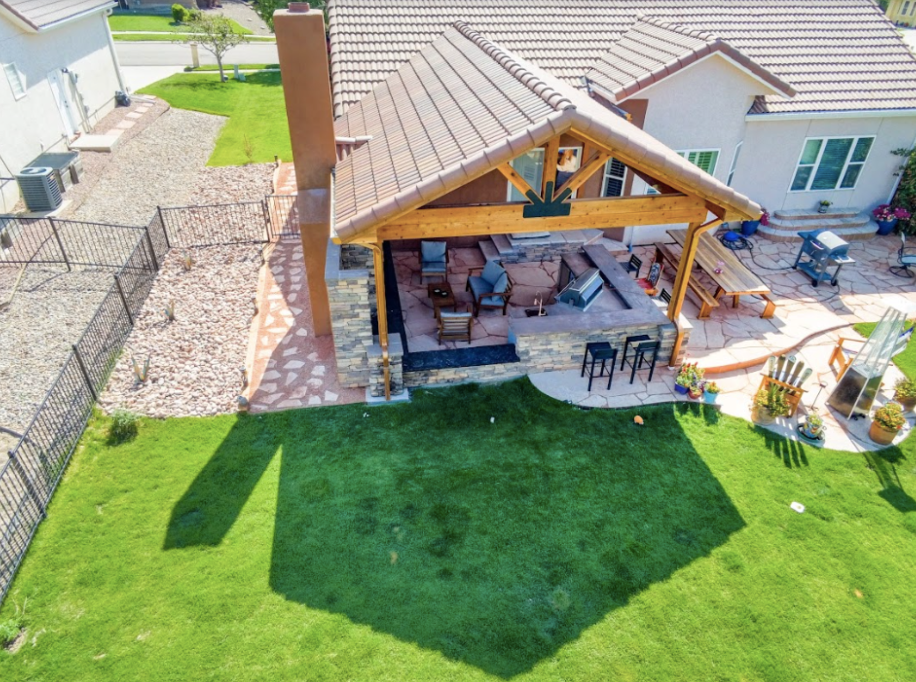 A backyard with a covered outdoor patio featuring wooden beams, stone flooring, and outdoor seating, adjacent to a well-manicured green lawn, enclosed by a black metal fence, with neighboring houses visible in the background.