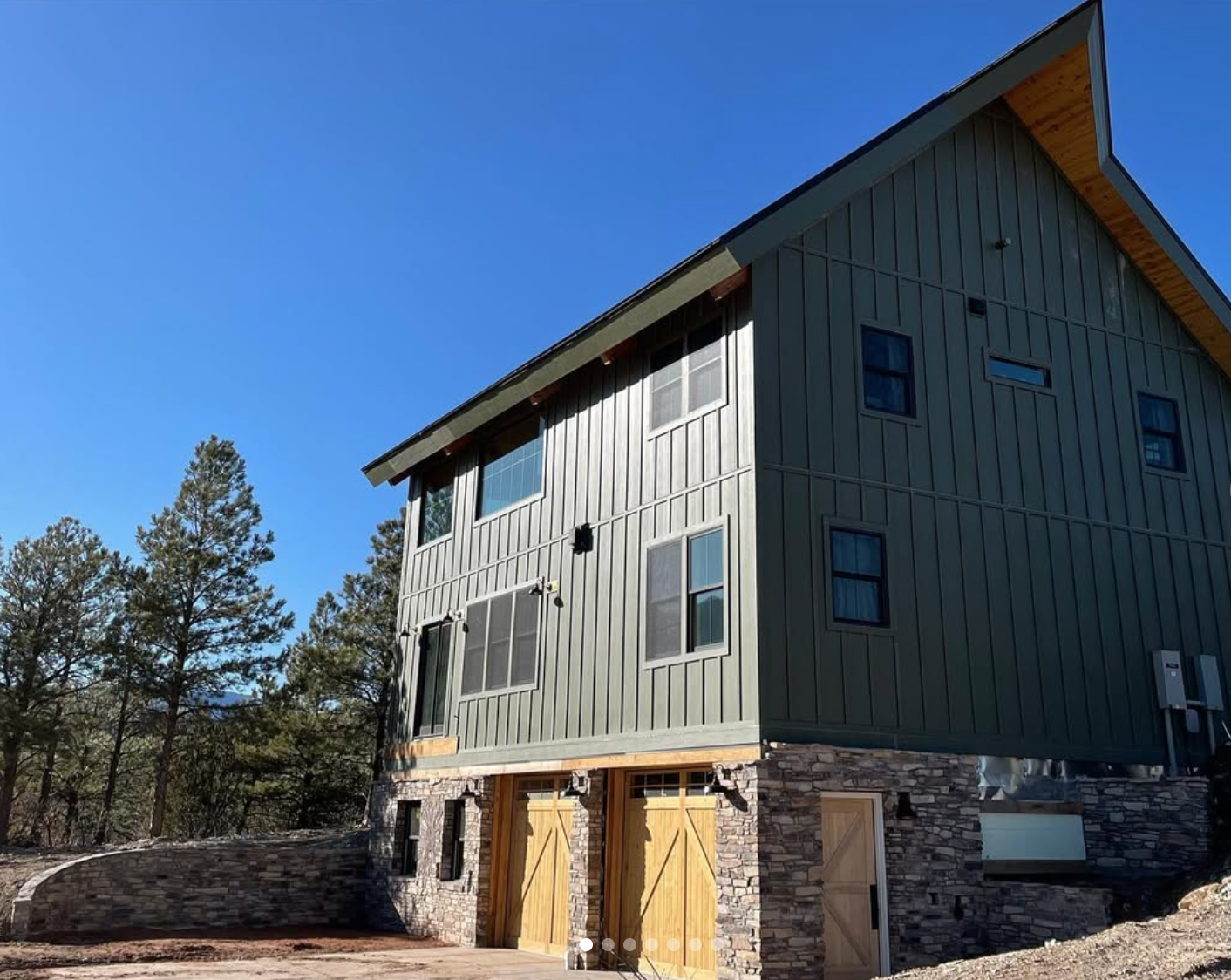 A modern house with green vertical siding, wooden accents, and a stone foundation, surrounded by trees under a clear blue sky.
