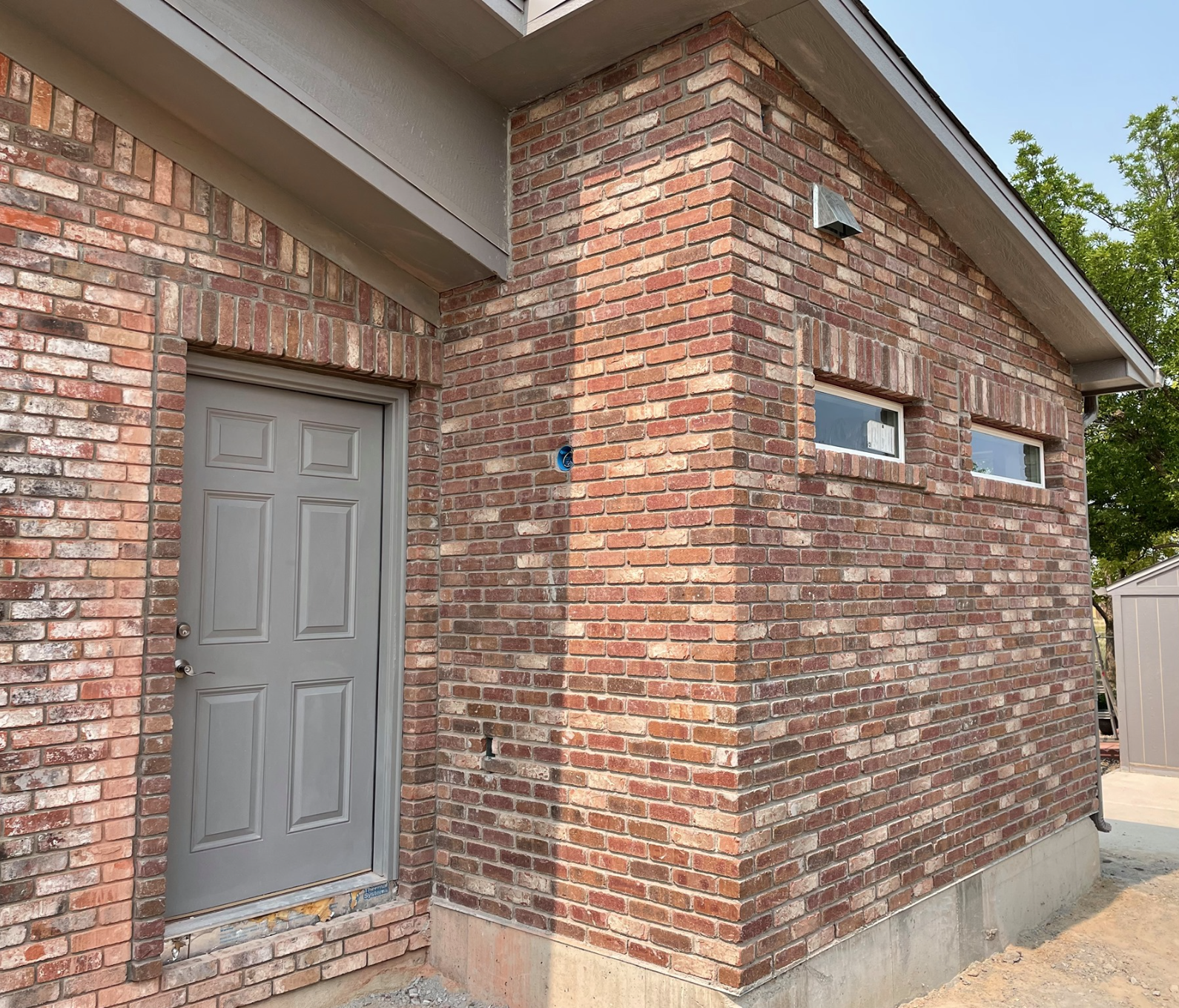 Brick exterior of a building with a grey door and two small horizontal windows, surrounded by trees.