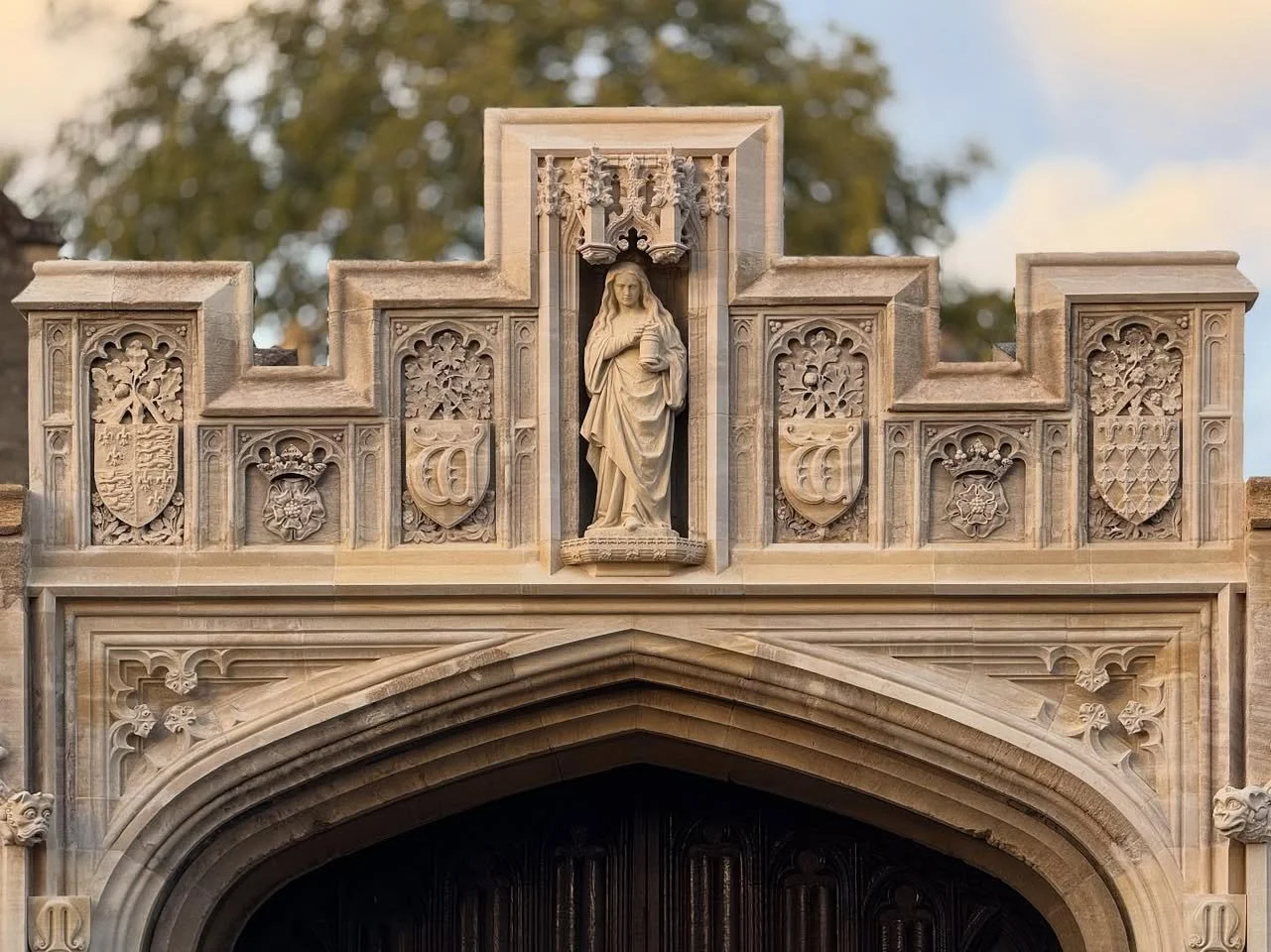 Gothic-style stone archway with detailed carvings and a central statue of a woman holding a lantern, set against a blurred background of trees and sky.