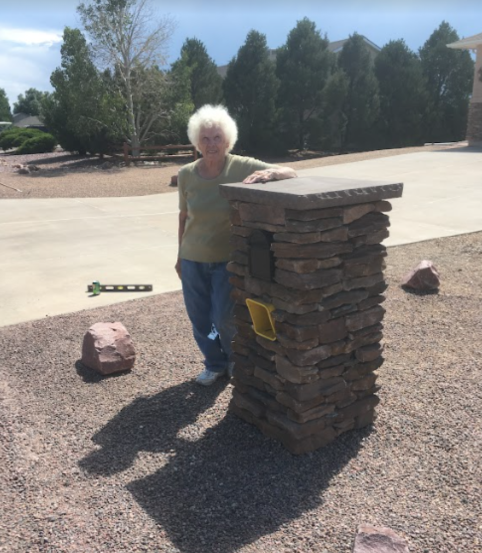 An elderly woman with white curly hair standing next to a tall stone chimney in a backyard. She is smiling and resting her hand on top of the chimney. There are large rocks and a level tool on the ground, and a concrete driveway with trees and a hous