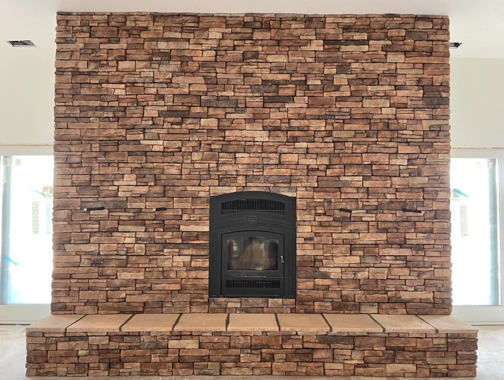 Interior view of a large brick fireplace with a black wood-burning stove inset, flanked by two windows, and a brick hearth in front.