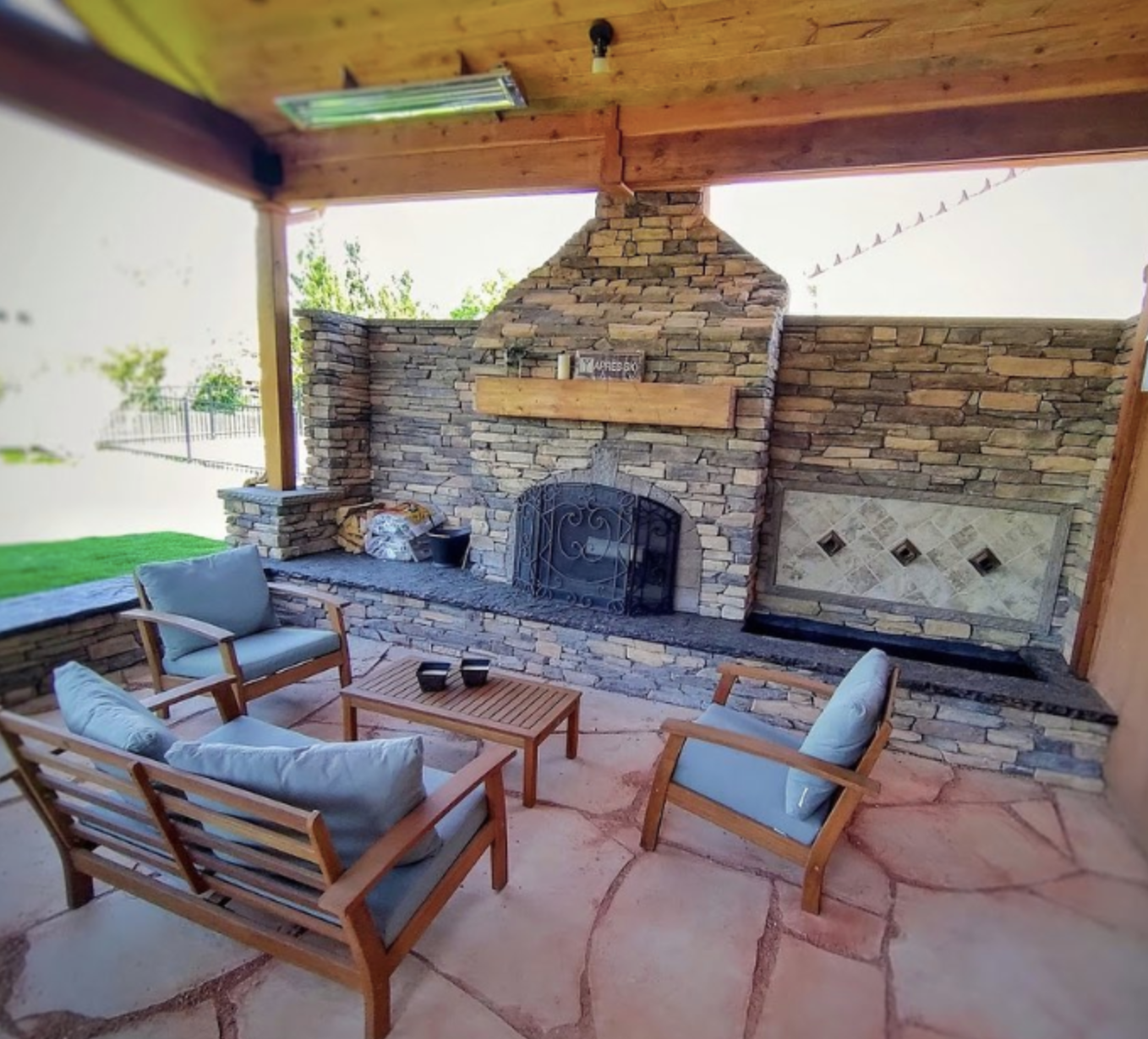 Outdoor patio with stone fireplace, wooden furniture with cushions, and a partial view of a backyard with green grass and a fence.