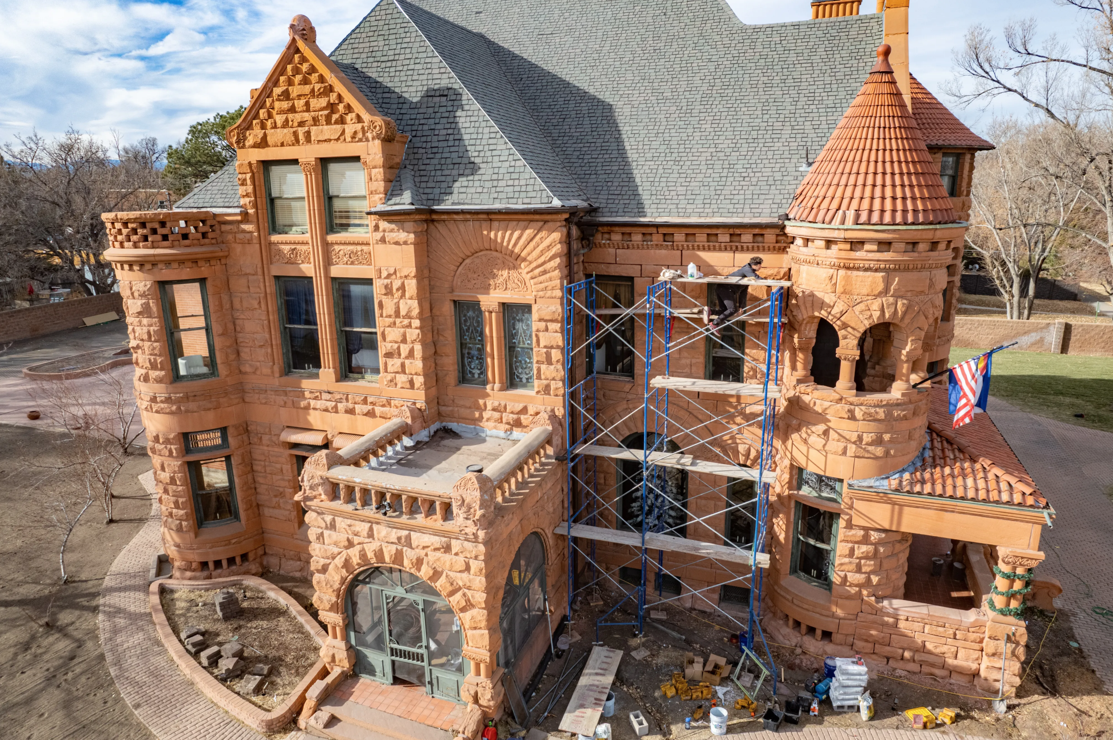 A large, historic-looking brick house under renovation with scaffolding on one side, workers, and an American flag hanging from the structure.