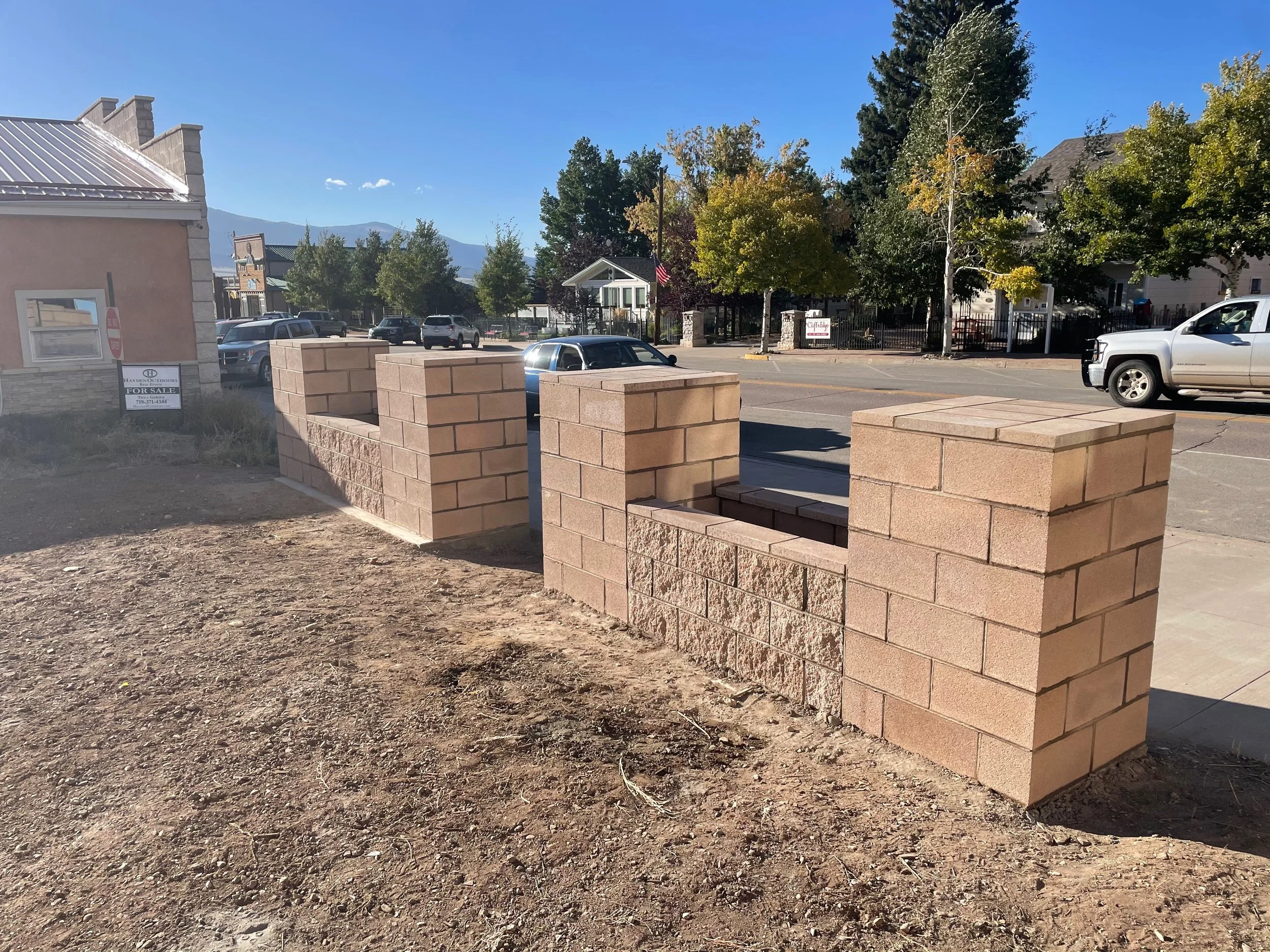 Construction of a brick wall with two open sections at a sidewalk, with parked cars, trees, and buildings in the background under a clear blue sky.