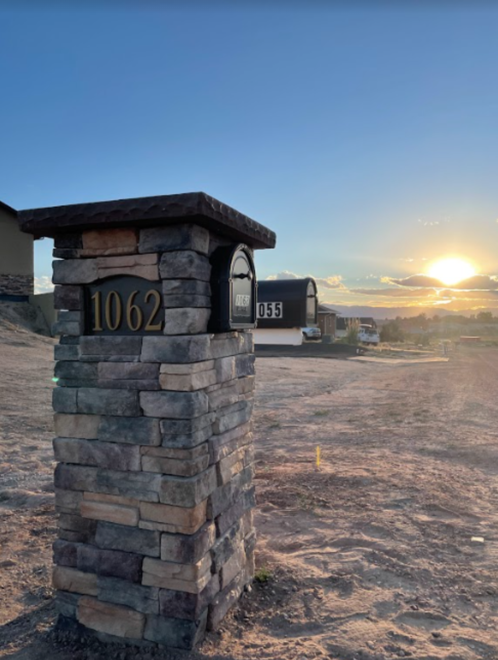 A stone mailbox with house number 1062 stands on a dirt lot during sunset, with other mailboxes and distant hills visible in the background.