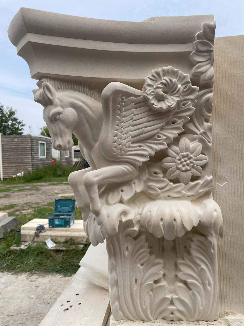 A detailed white stone or sculpted decoration featuring a horse with wings, surrounded by flowers and ornate patterns, set outdoors with a partly cloudy sky and temporary structures in the background.
