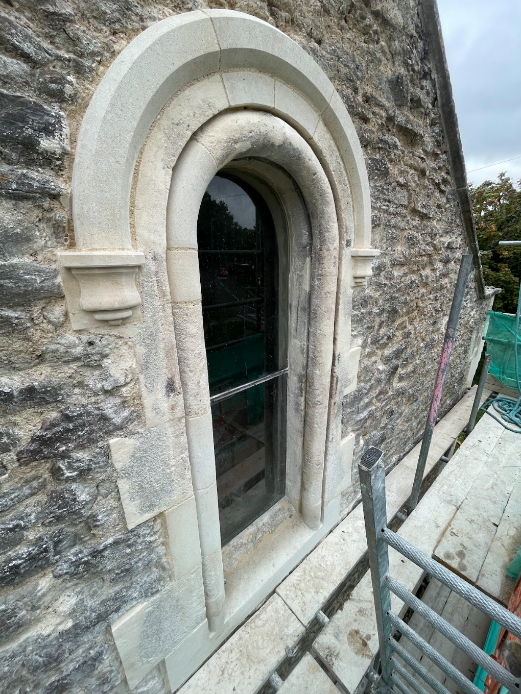 A close-up of a stone arch window on a stone building under renovation, with scaffolding and construction tools visible around it.