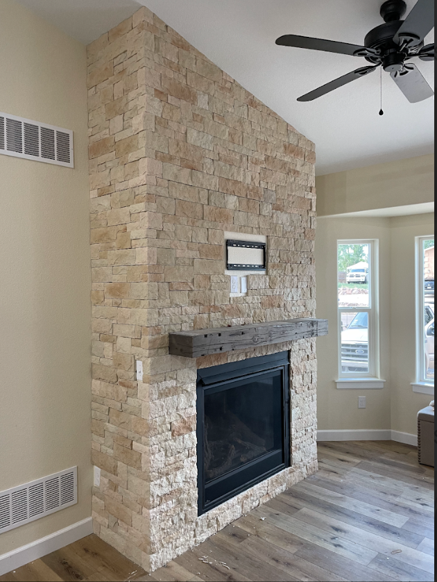 Living room corner with a stone fireplace, gray wooden mantel, and a ceiling fan; windows overlooking a patio with parked cars.