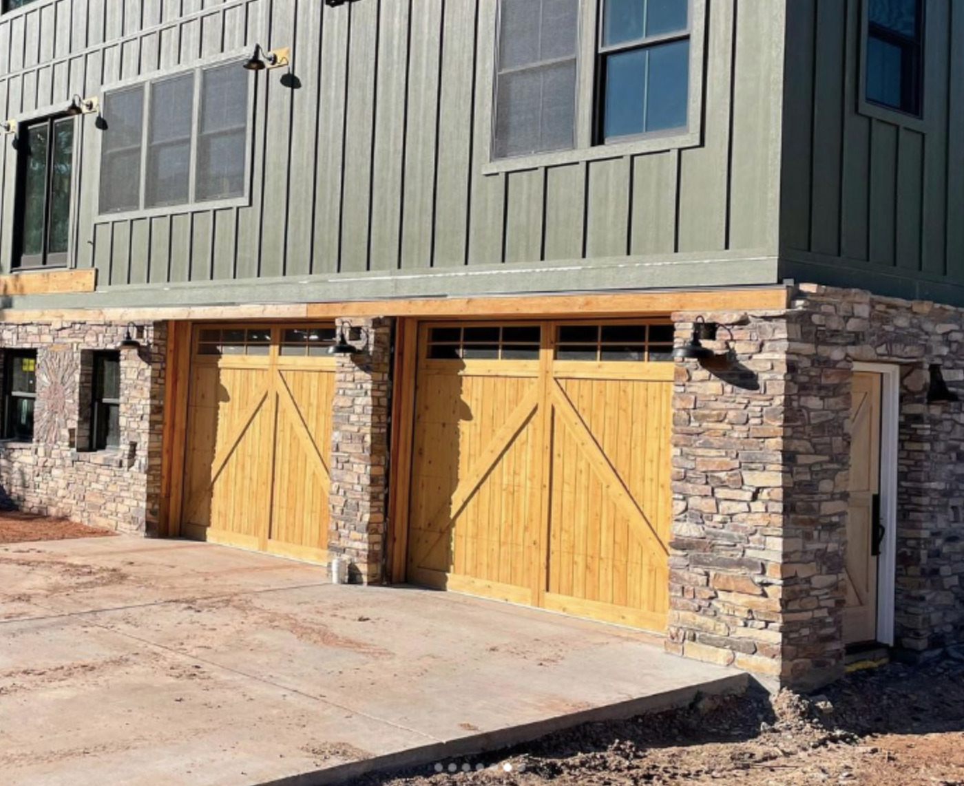 Newly constructed building with a stone and wood facade, featuring two large wooden garage doors with a diagonal cross design, and black exterior wall lights installed above the doors.