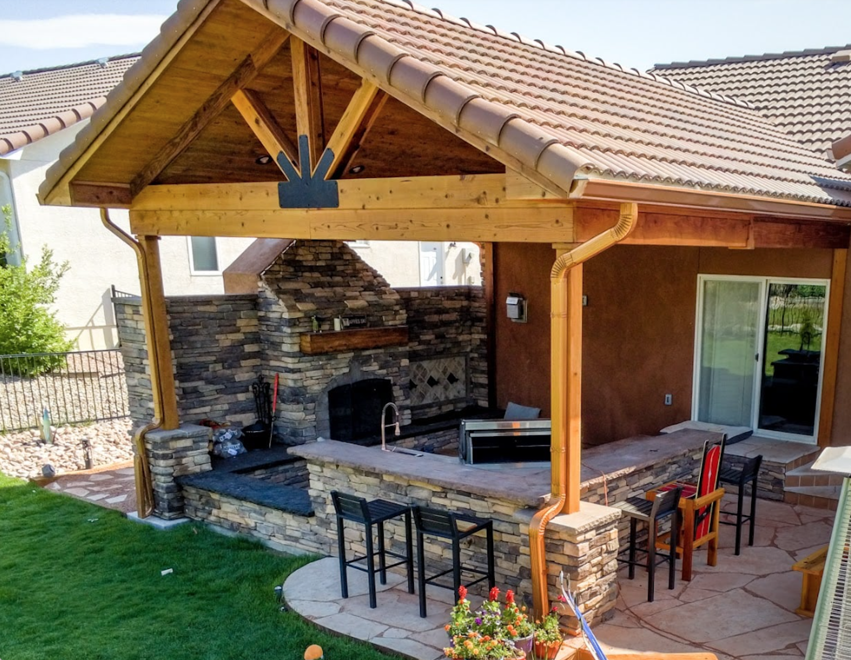 A backyard patio with a stone outdoor fireplace and grill, a stone countertop bar area with chairs, and a wooden-covered roof structure over the patio.