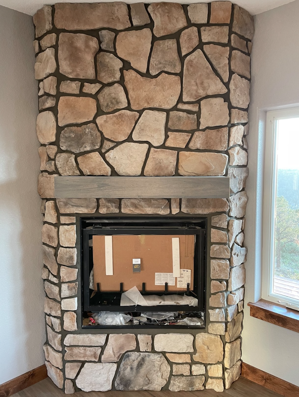 A stone fireplace built into a wall, with a wooden mantel and a window to the right, showing the interior construction of the fireplace with packaging materials inside.