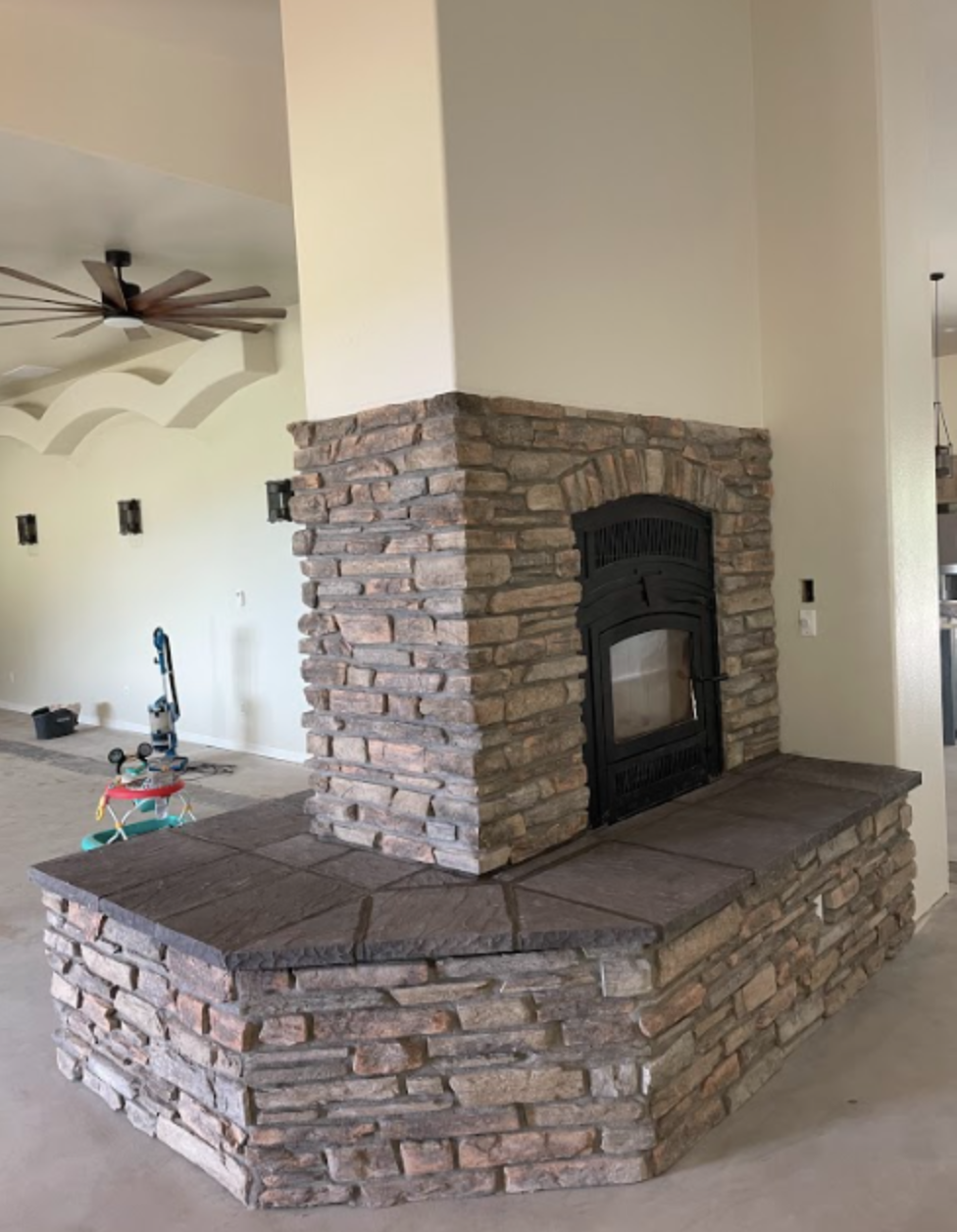 Living room with a brick fireplace, wooden ceiling fan, and construction tools in the background.
