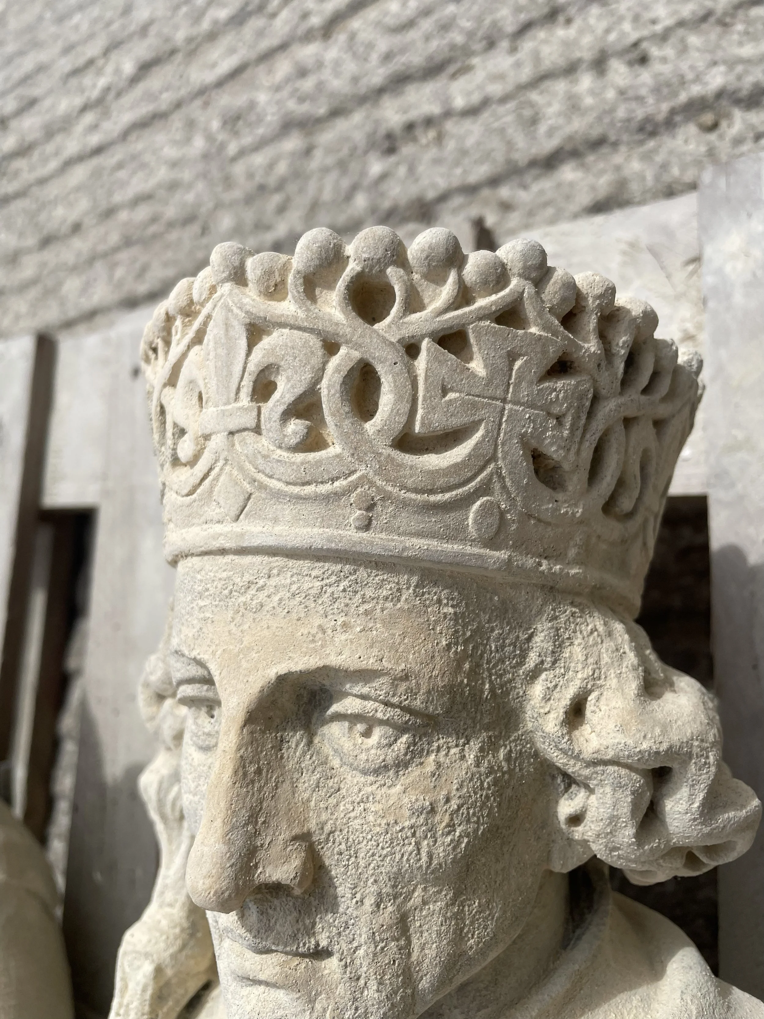 Close-up of an ancient carved stone bust of a person wearing a detailed crown or headdress, with a neutral facial expression, set against a stone wall background.