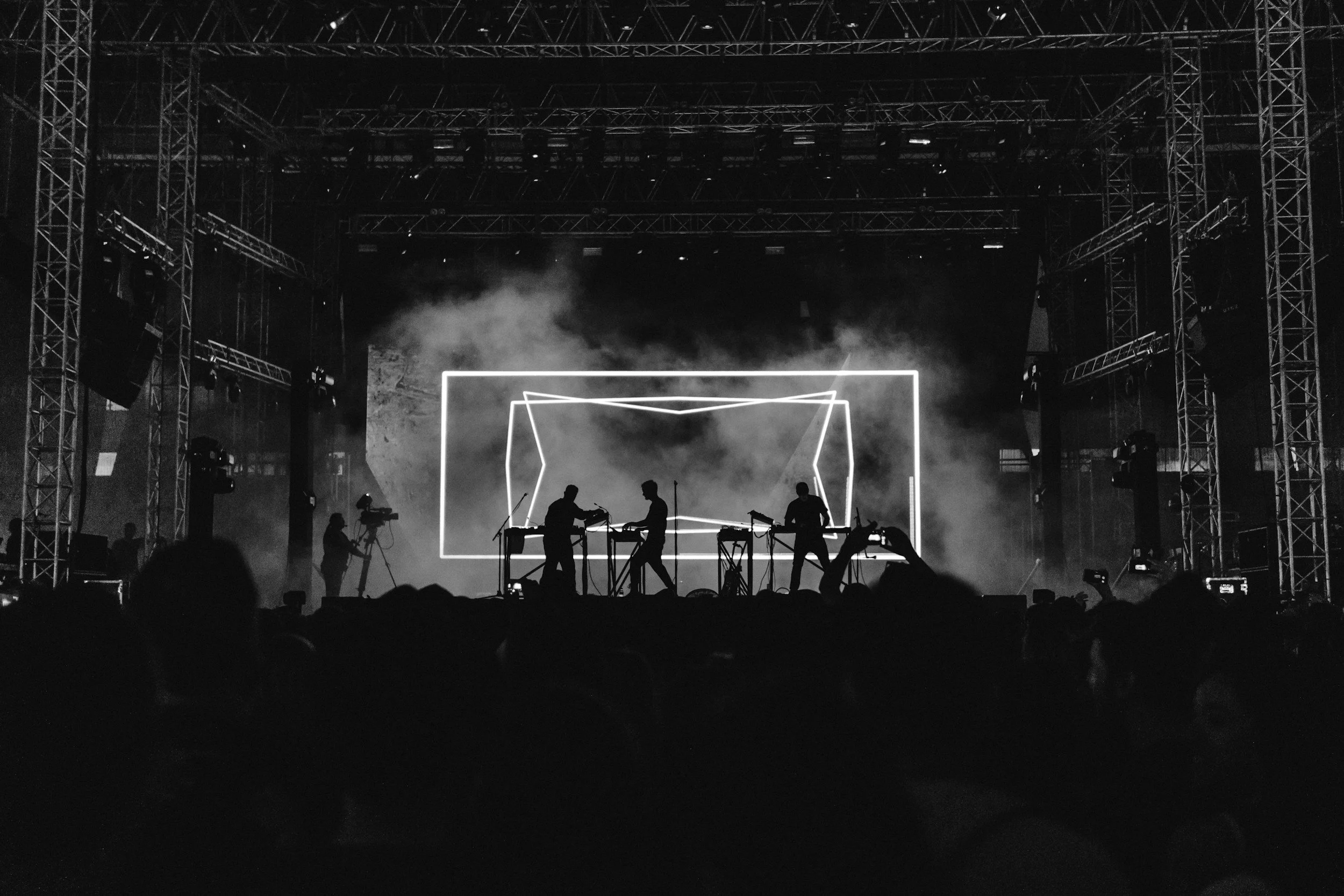A live music concert with a stage, performers playing instruments, and a large digital screen displaying geometric shapes, with audience silhouettes and stage lighting.