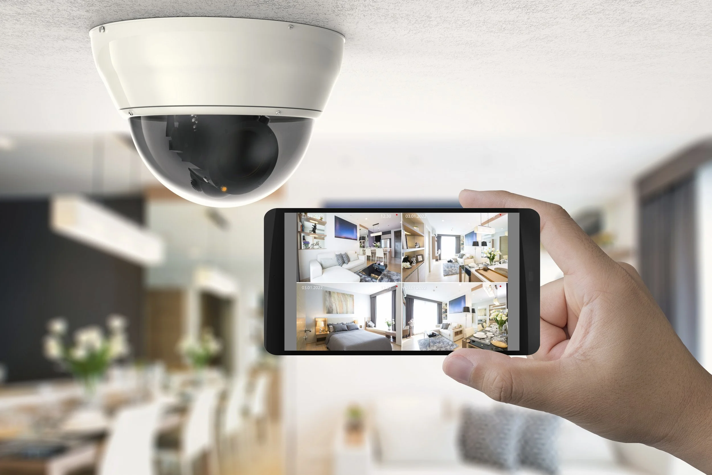Person holding a smartphone monitoring multiple security camera feeds inside a modern, well-lit living room.