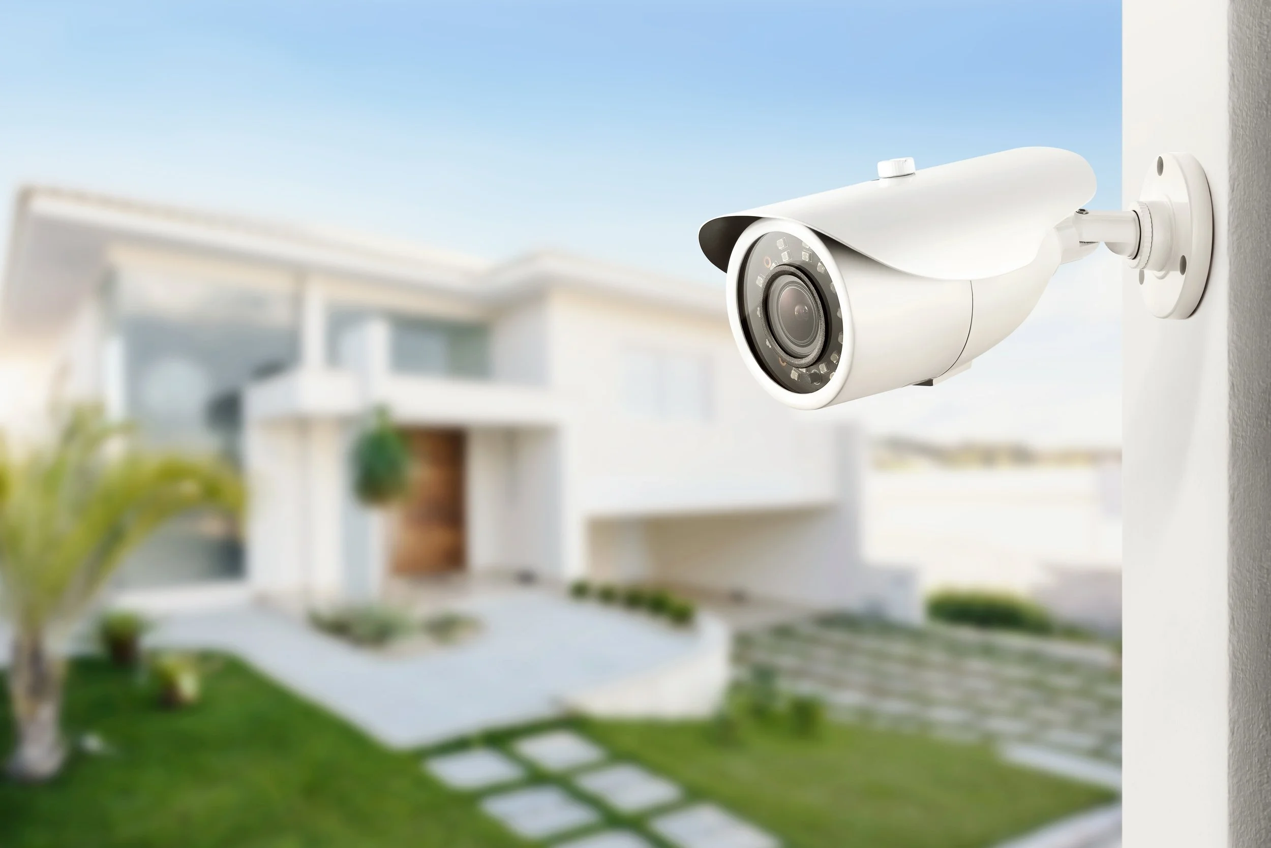 White security camera mounted on exterior wall of modern house with front yard, pathway, and lawn, blurry background.