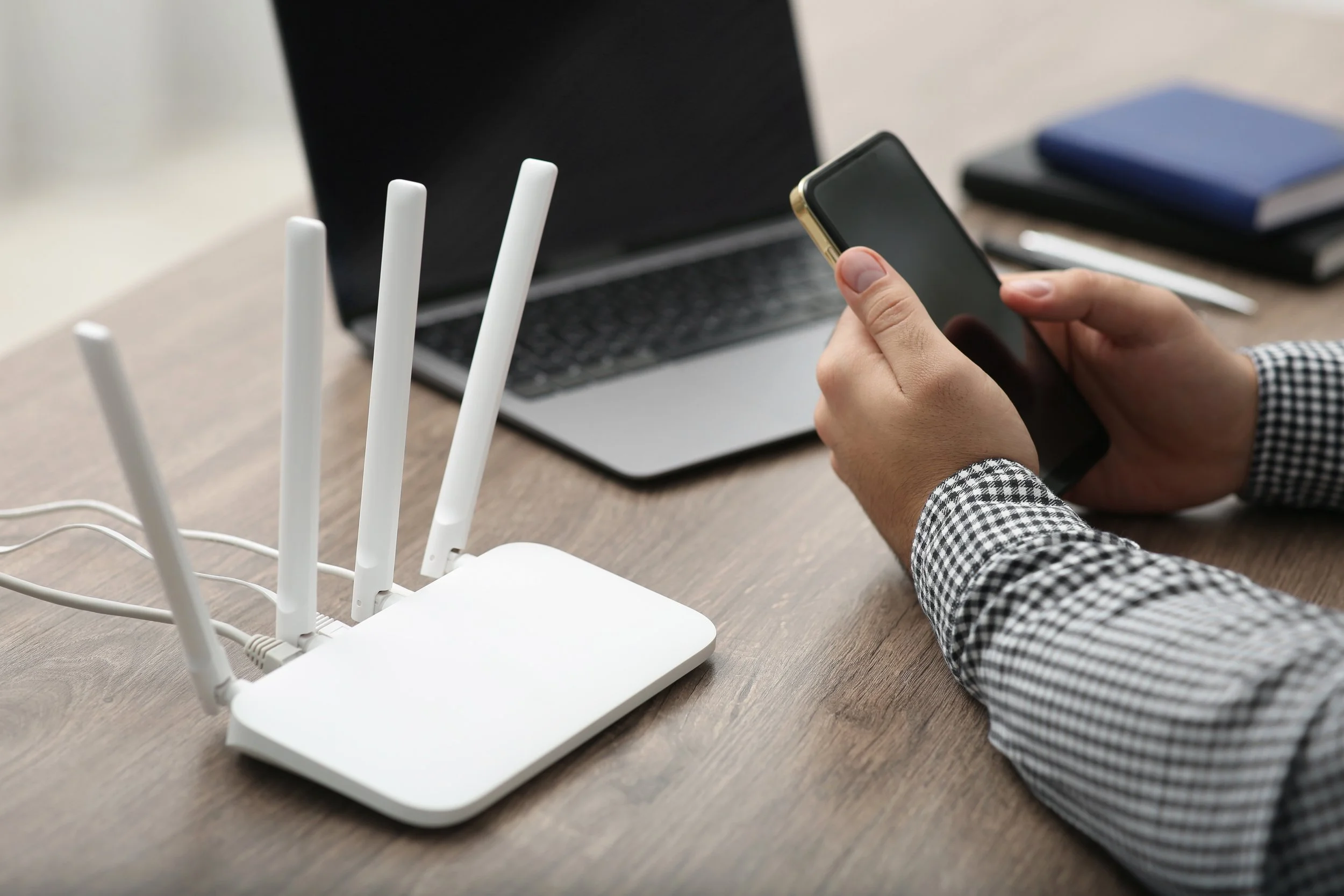 Person in checkered shirt using smartphone with Wi-Fi router on wooden desk, open laptop, notebooks, and pen in background.