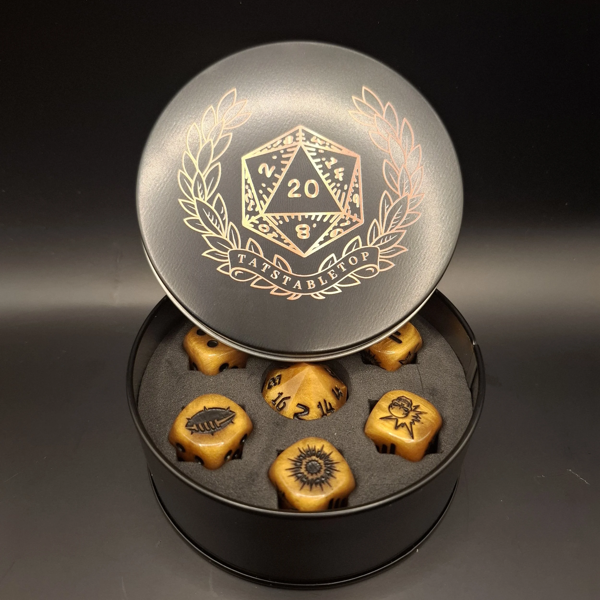 A round black container with a removable lid featuring a 20-sided die design and the words "Tatstabelle Top". Inside are six custom-shaped dice with symbols on each face.