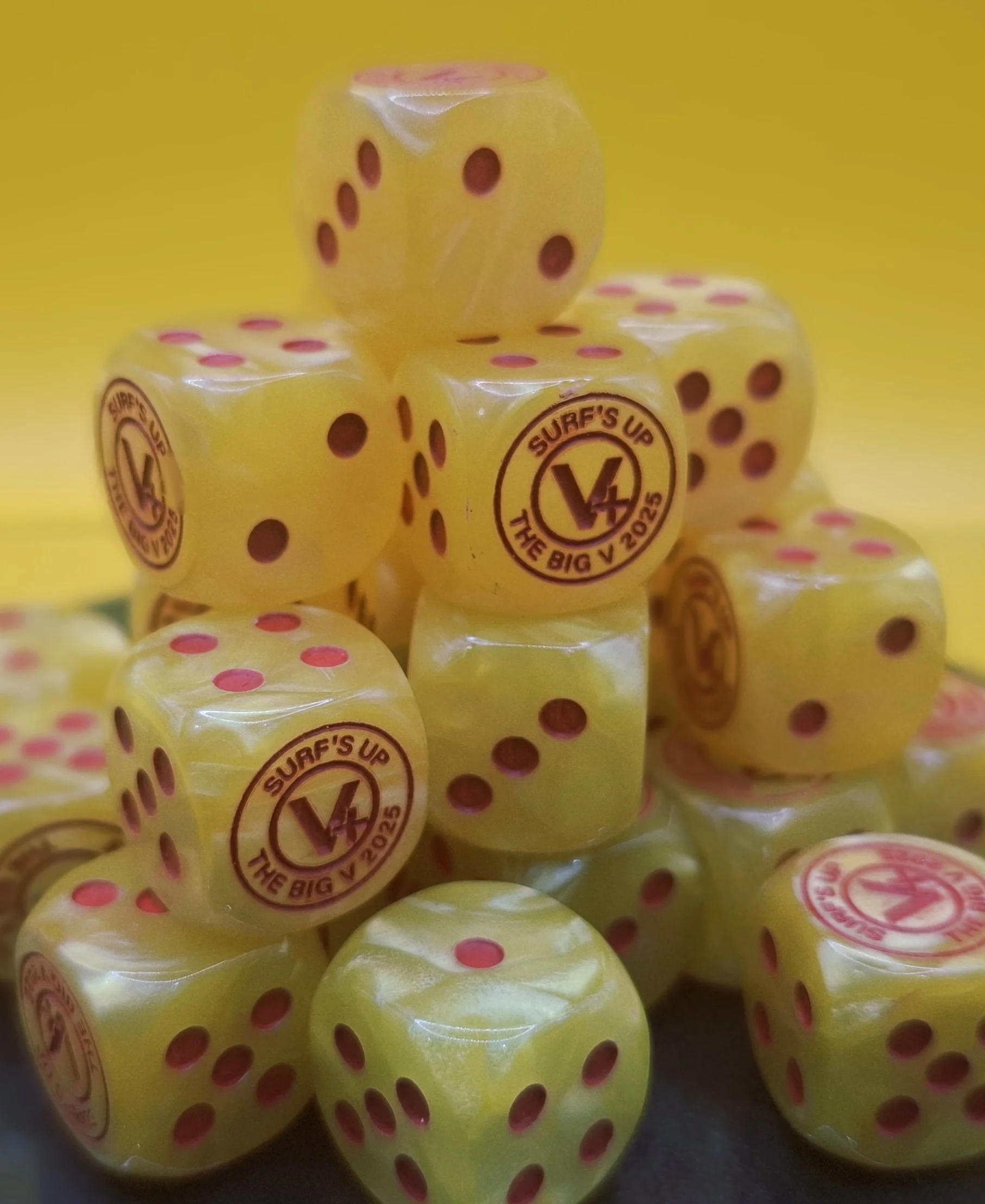 A pile of yellow dice with pink pips and a logo reading 'Surf's Up The Big V 2025' on some of the dice.