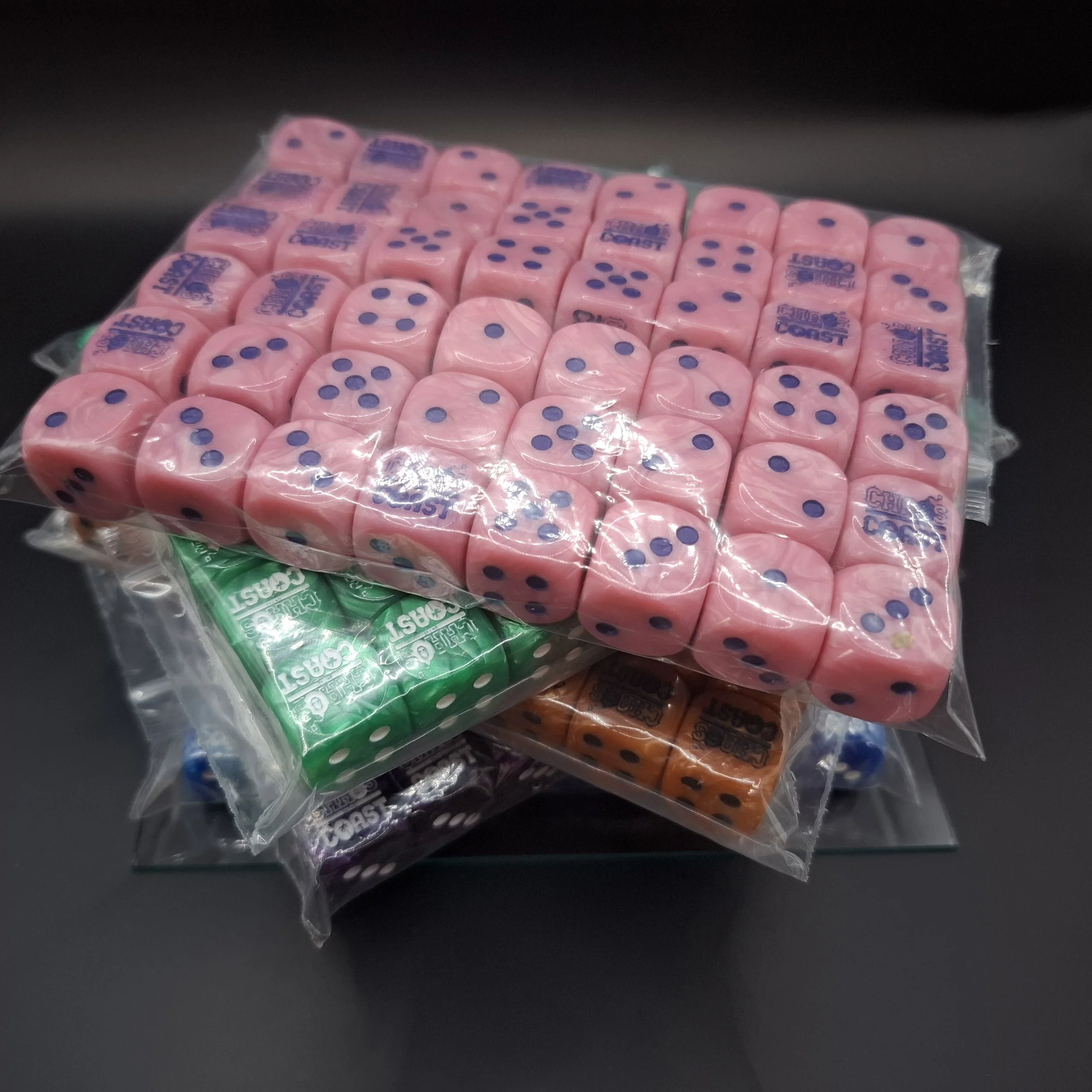 Collection of colorful dice in plastic bags, including pink, green, purple, and orange, on a black surface.