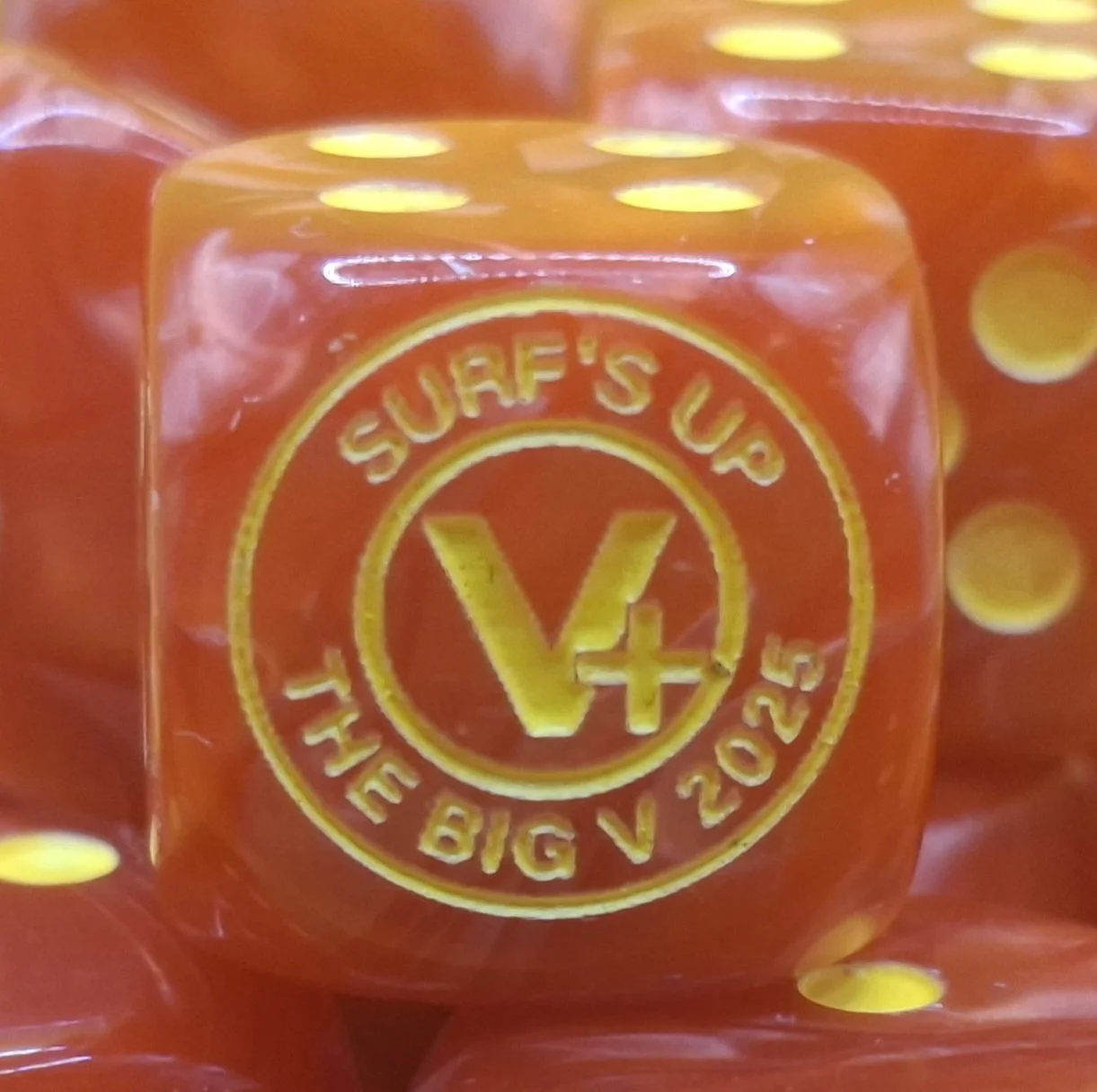 Close-up of a red dice with yellow dots and yellow embossed text and logo reading 'SURF'S UP THE BIG V 2025'.