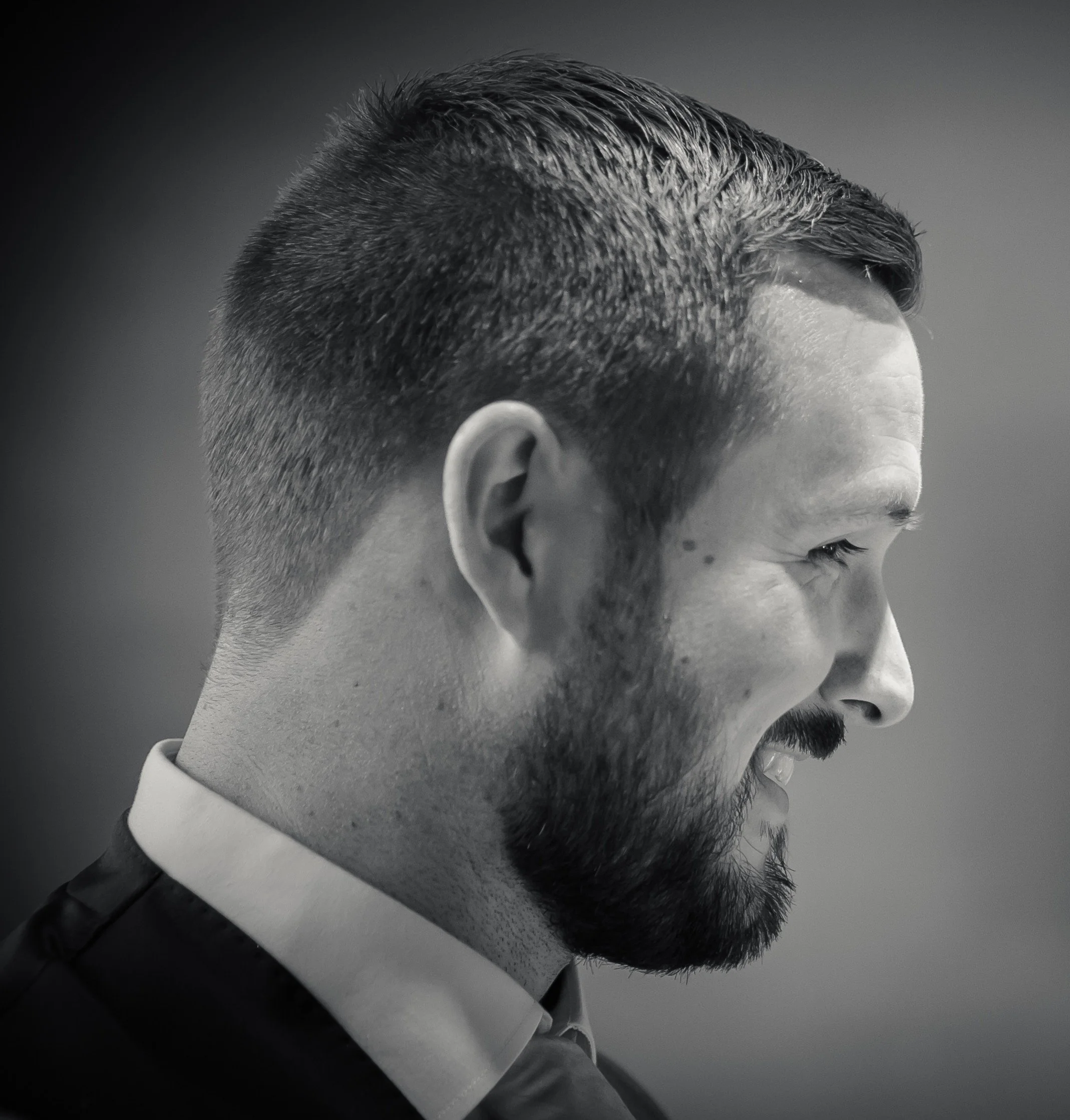 Side profile of a smiling man with short, styled hair and a beard in black and white.