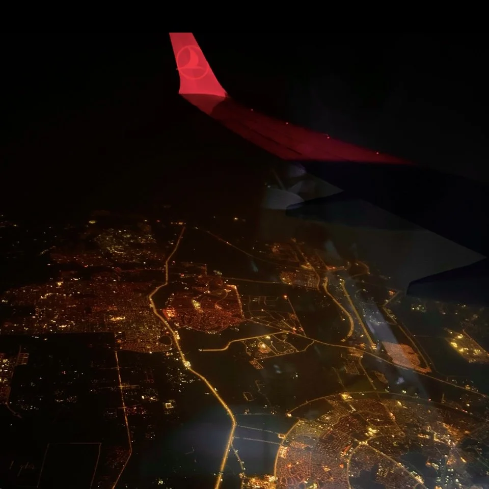 Nighttime aerial view of a city seen from an airplane window, with illuminated streets and buildings, and wing of the airplane visible in the upper part of the image.