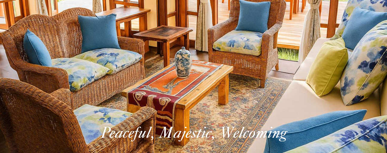 A cozy living room with wicker and fabric sofas, colorful pillows, a wooden coffee table with a decorative vase, a patterned rug, and large windows with curtains letting in natural light.