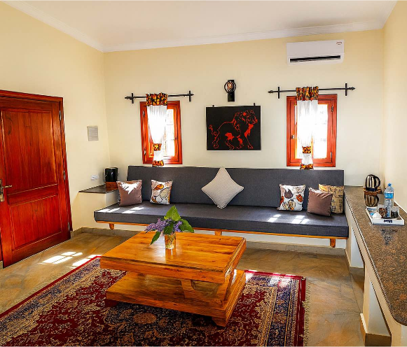 Living room with a wooden door, beige walls, a gray couch with multiple throw pillows, a wooden coffee table with a plant, a colorful rug, two small windows with curtains, and framed art on the wall.