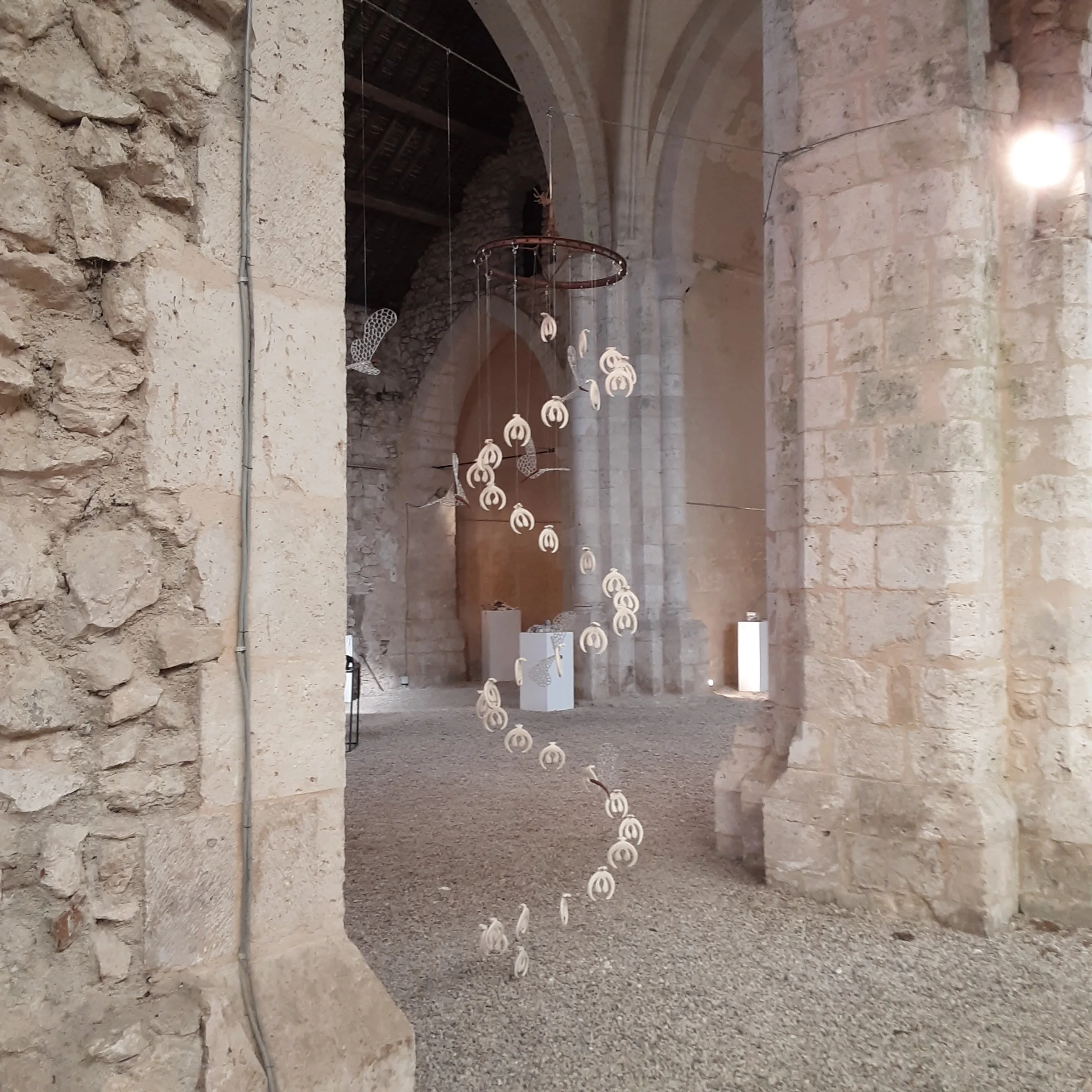 Une installation artistique suspendue dans un bâtiment en pierre, composée de plusieurs formes pendantes en forme de spirale et de vol d'oiseaux, avec des arches et murs en pierre en arrière-plan.