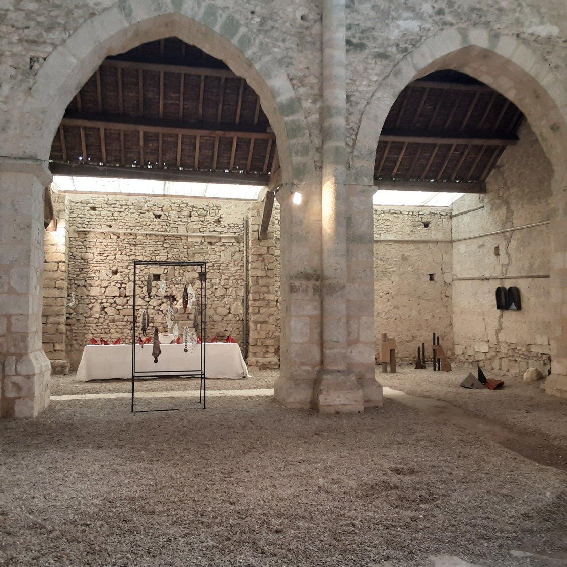 Une salle ancienne en pierre avec des arches et un mur en briques, une table décorée de vaisselle, un porte-manteau avec des objets suspendus, et divers objets en catimini sur le sol.