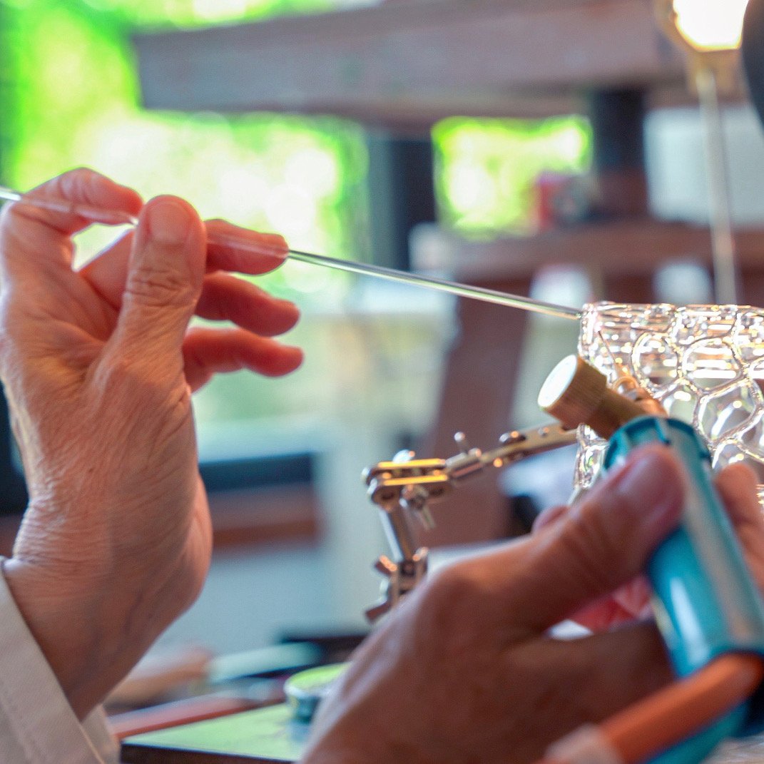 Personne en train de souder une baguette en verre, avec un chalumeau, dans un atelier lumineux.