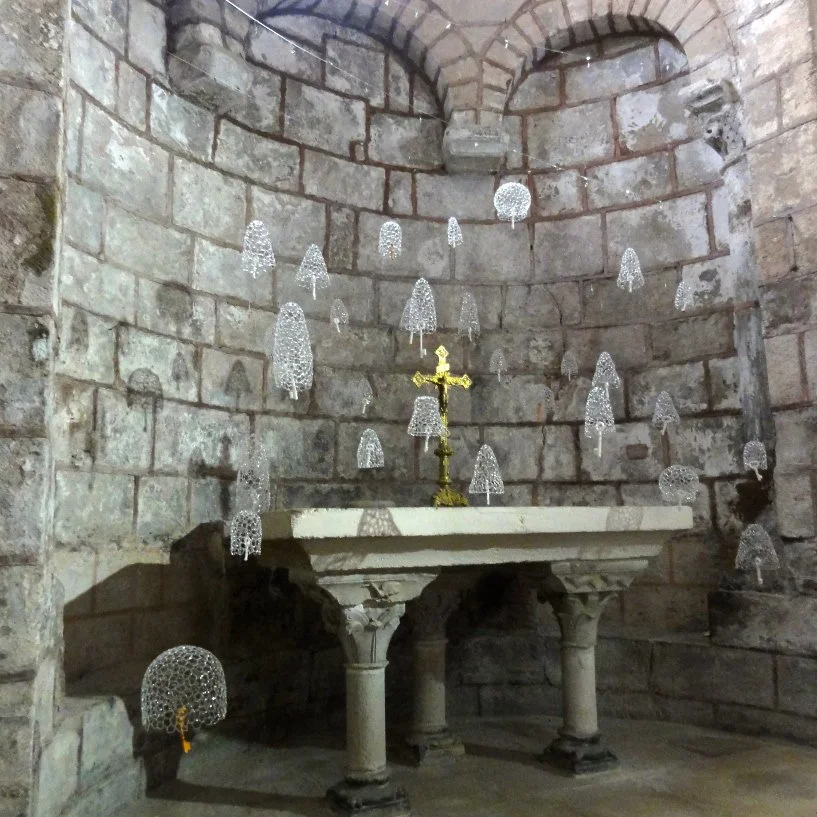 Une ancienne chapelle en pierre avec un autel en pierre, une croix en métal doré au centre, et des cloches de verre transparent dont les battants sont en céramique ou verre  en forme de clés, suspendues au plafond.