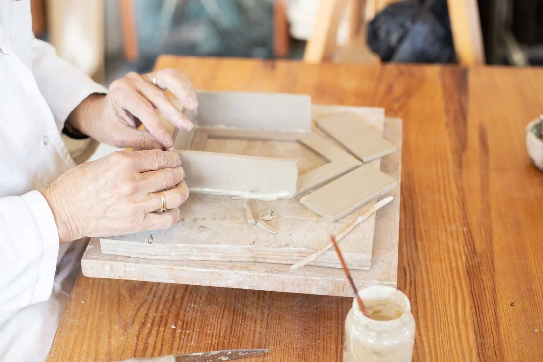 Une personne façonne de la terre disposée sur un plateau en bois, avec des outils, dans un atelier.