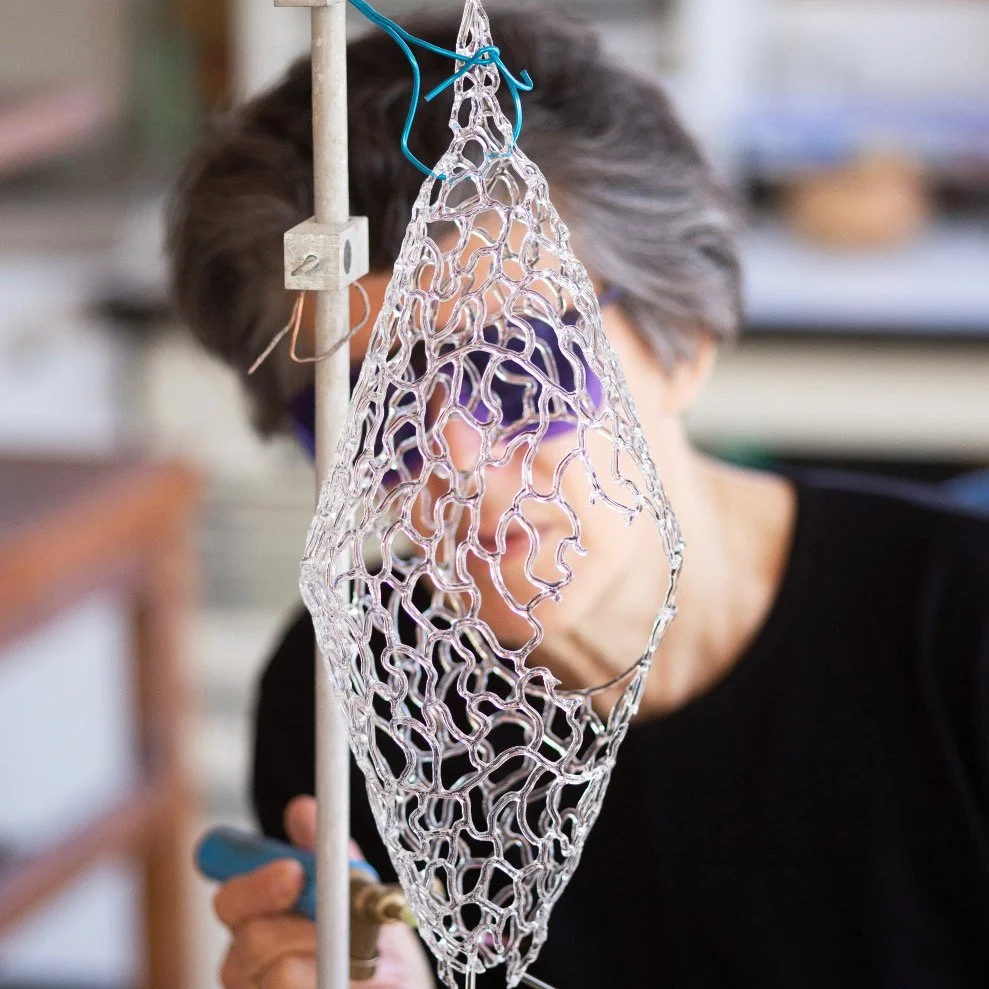 Une personne travaillant sur une sculpture en verre complexe, suspendue à un support, dans un atelier.