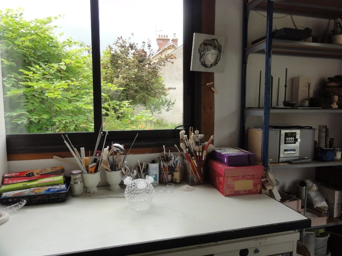 Un atelier d'art avec une table claire près d'une fenêtre donnant sur un jardin vert. Sur la table, il y a des pinceaux, des contenants, et un objet en verre.
