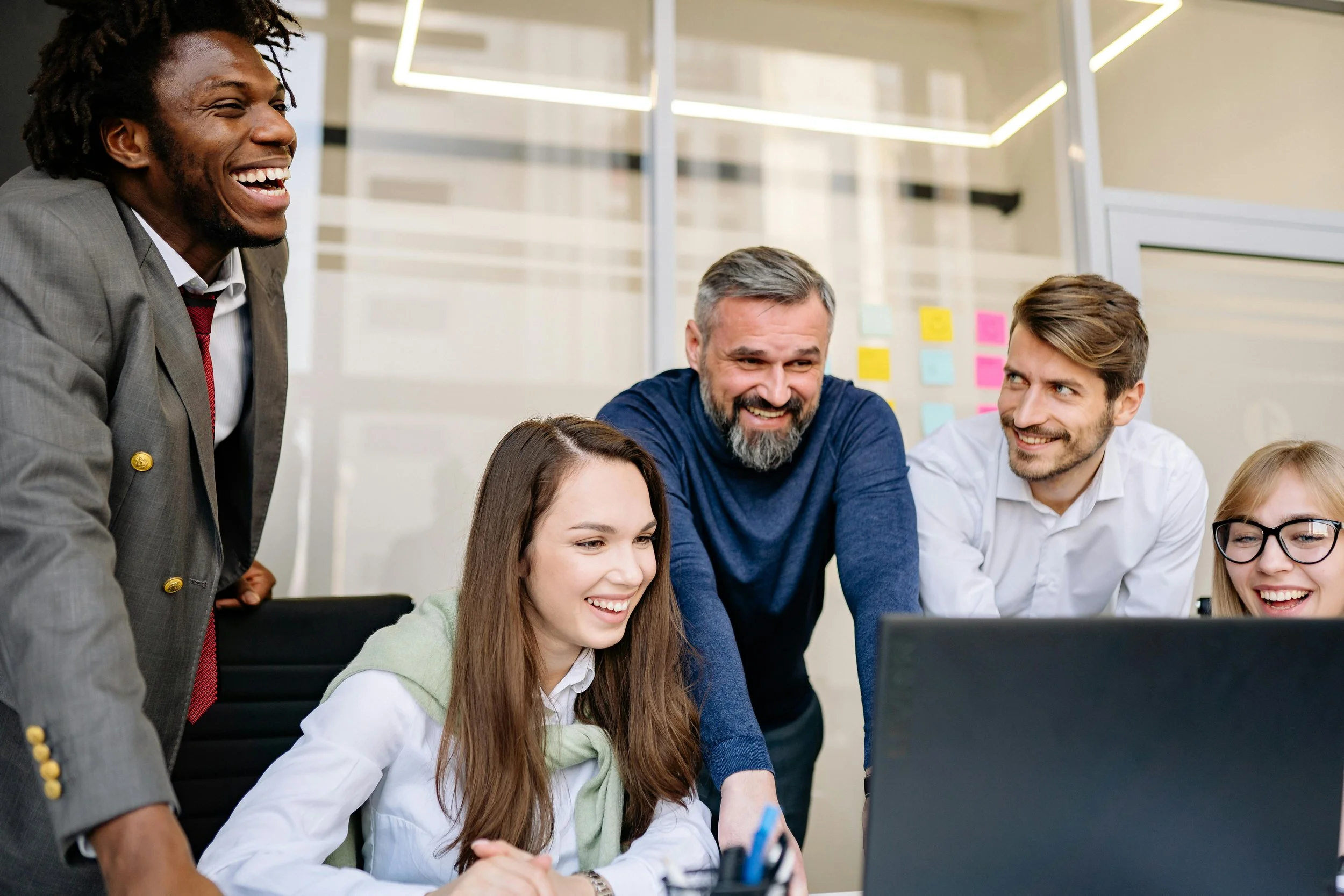 A group of five diverse colleagues gathered around a laptop, smiling, and engaging in a discussion in a modern office with glass walls and colorful sticky notes in the background.