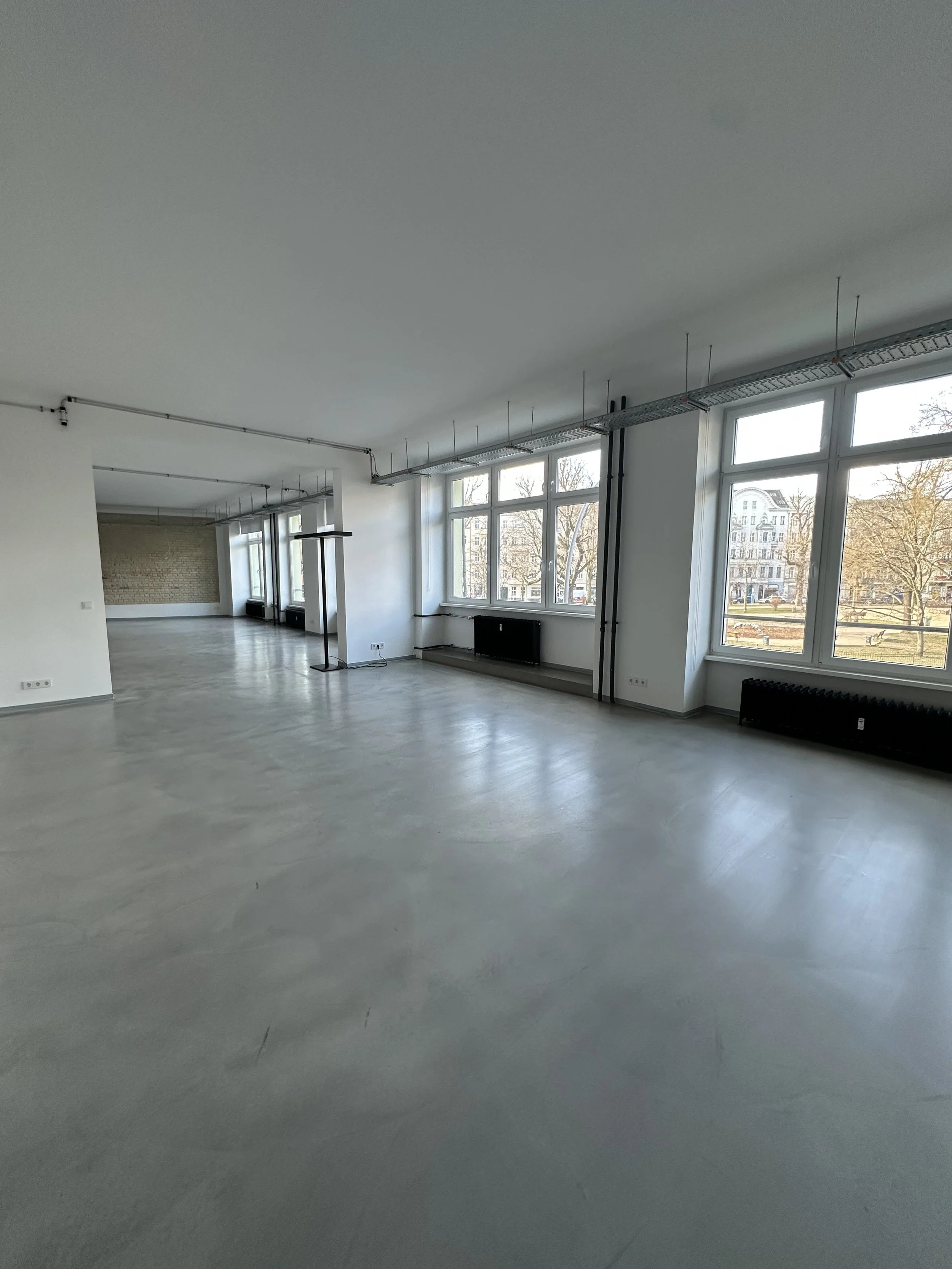 An empty, spacious room with large windows and a polished concrete floor, with visible ceiling wires and radiators beneath the windows.