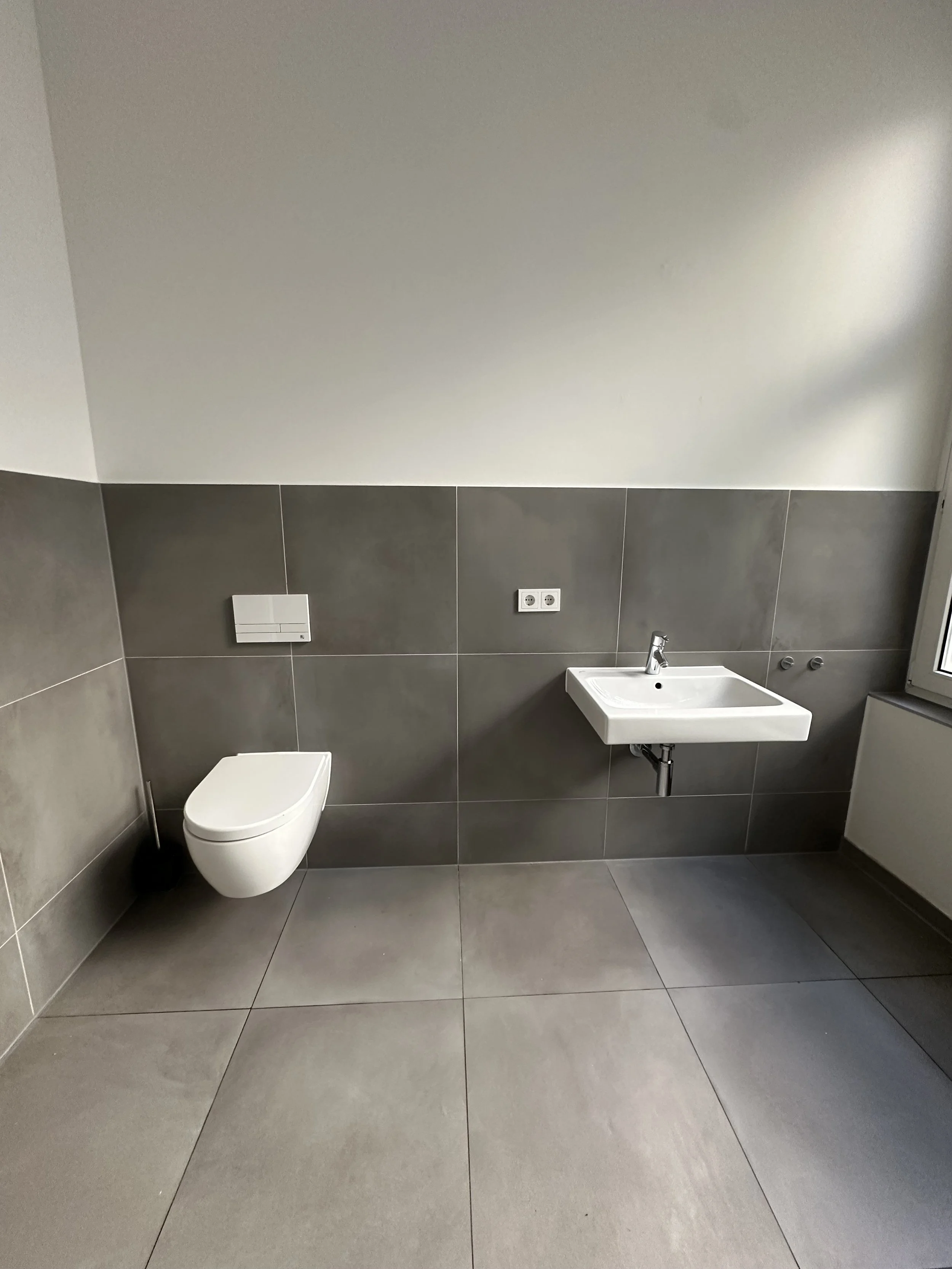 Empty bathroom with large gray tiles on the floor and walls, a small white wall-mounted toilet, and a white wall-mounted sink, near a window.