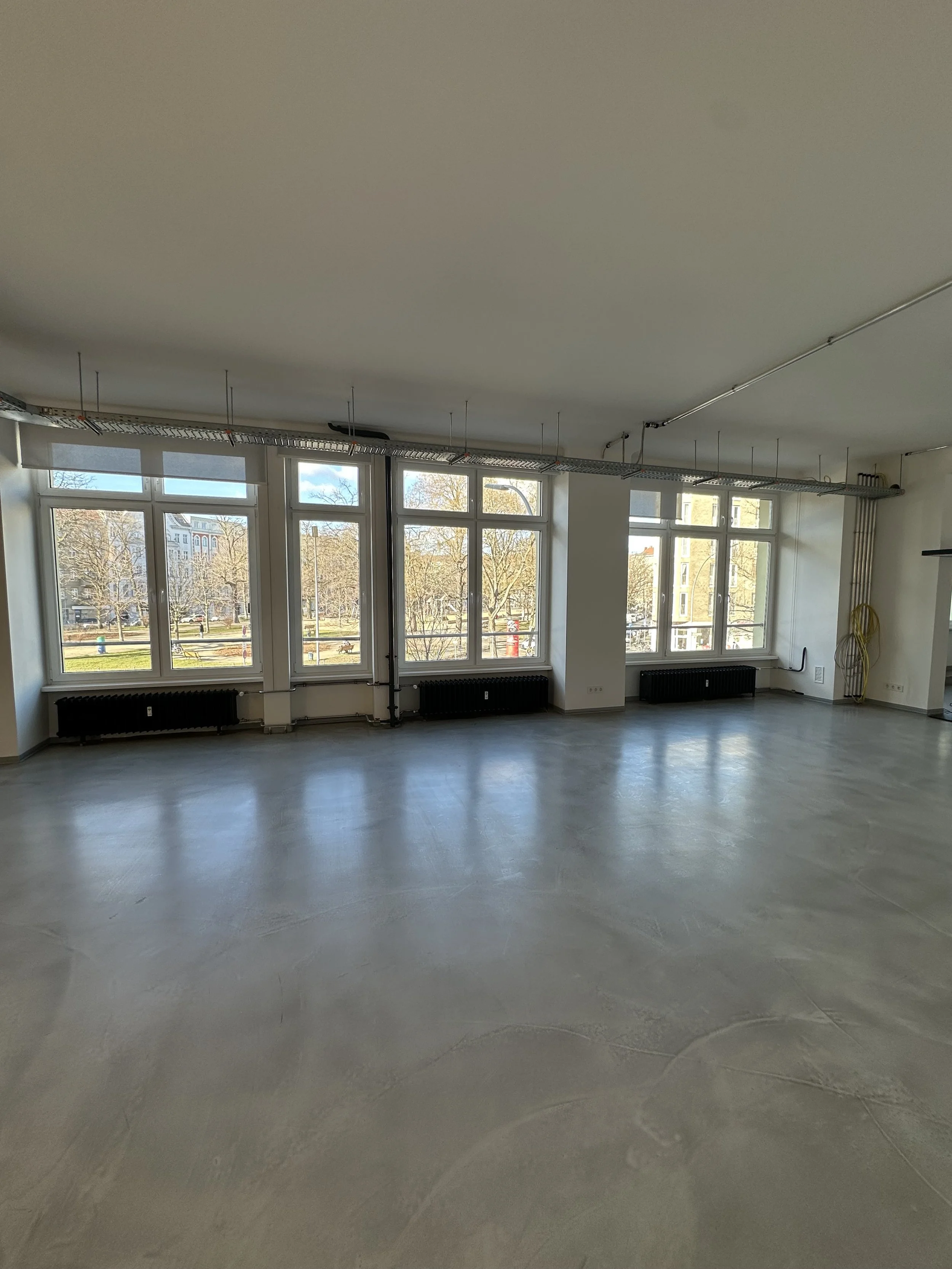 Empty office space with large windows, concrete floor, and some visible electrical wiring on the ceiling.