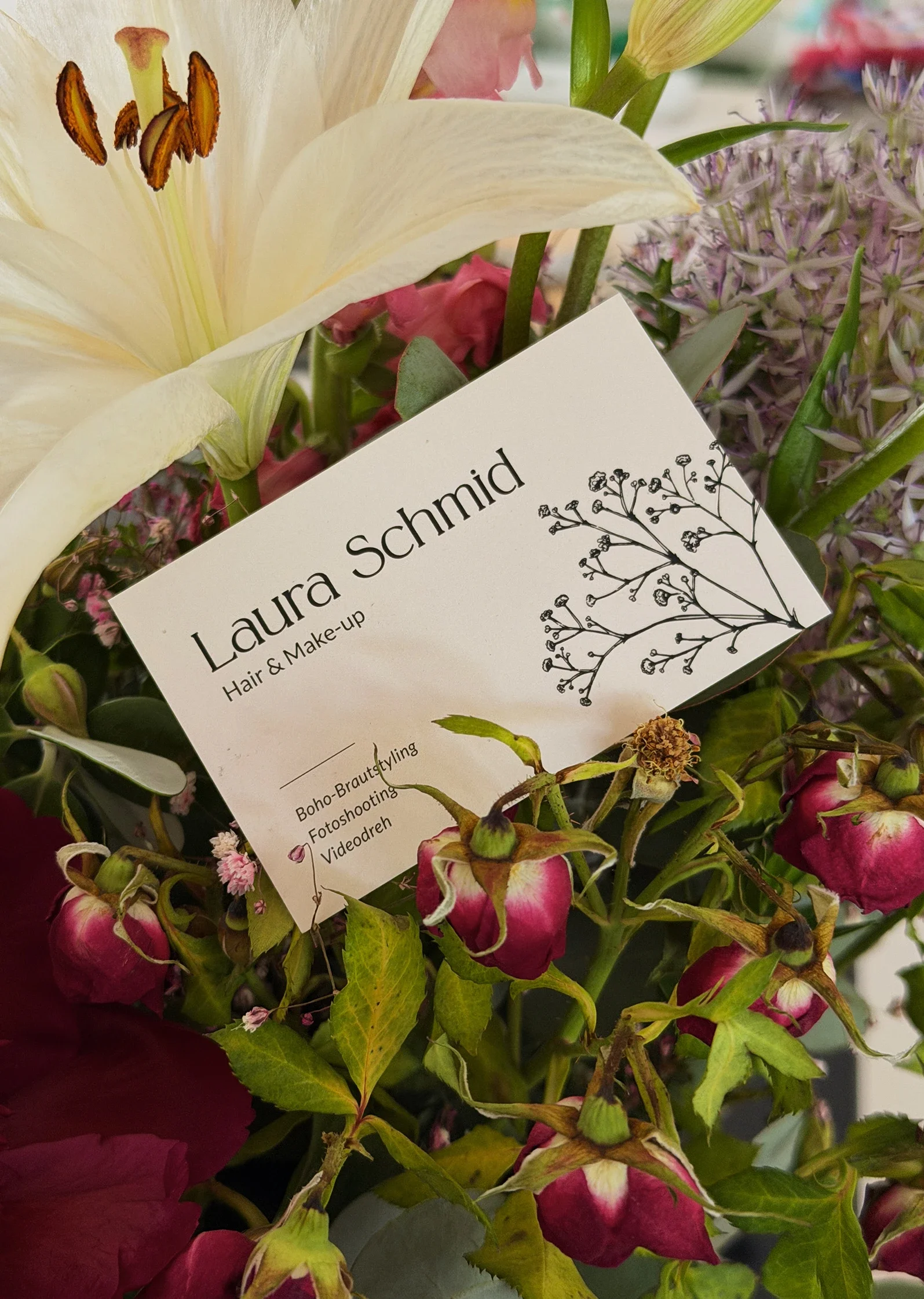 Zwischen Blumen liegt eine hübsch gestaltete Visitenkarte einer Hair und Make-up Artistin.
